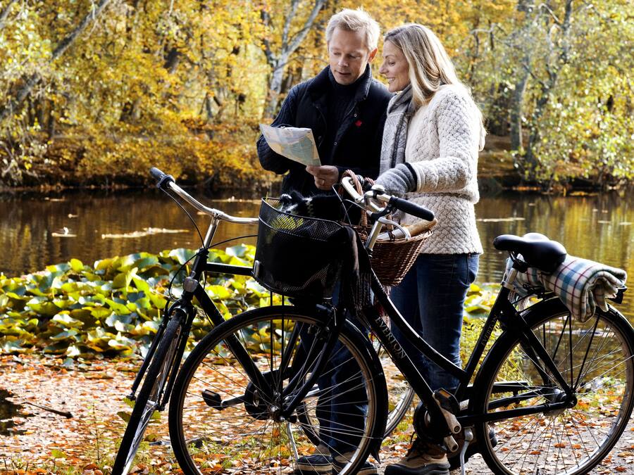 A couple standing by a bicycle with a map, near a body of water surrounded by autumn foliage.
