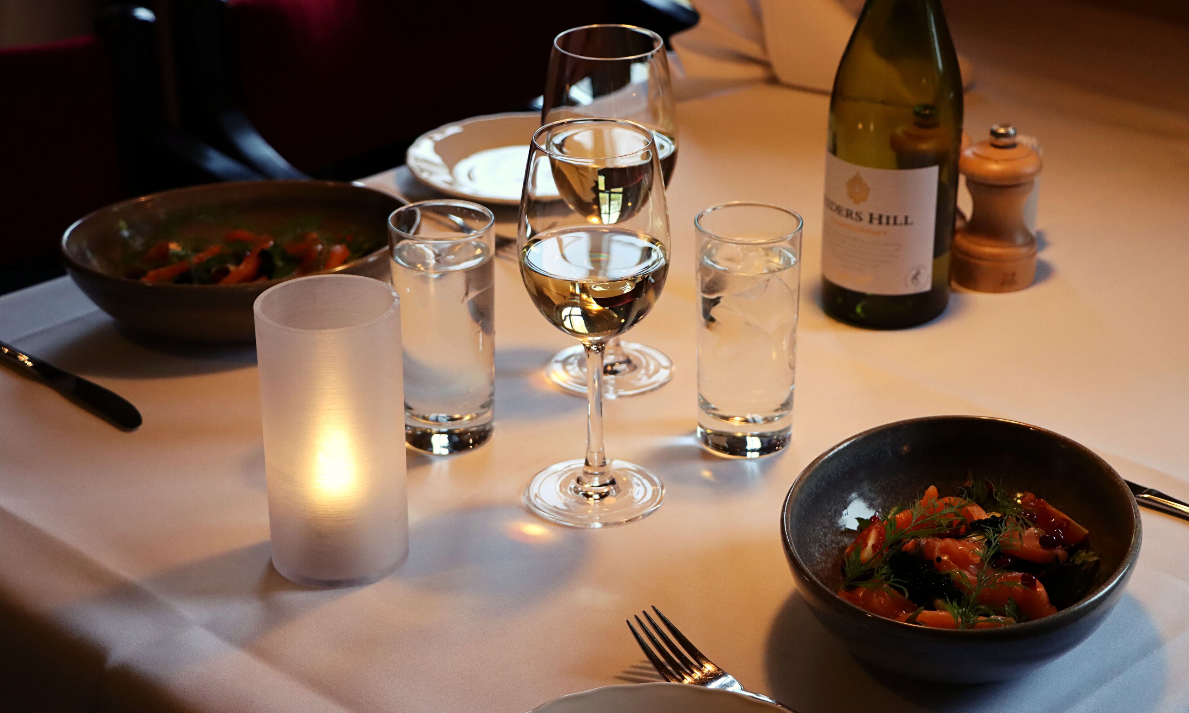 Una mesa de comedor con una vela, copas de vino, una botella de vino y un bol de comida.