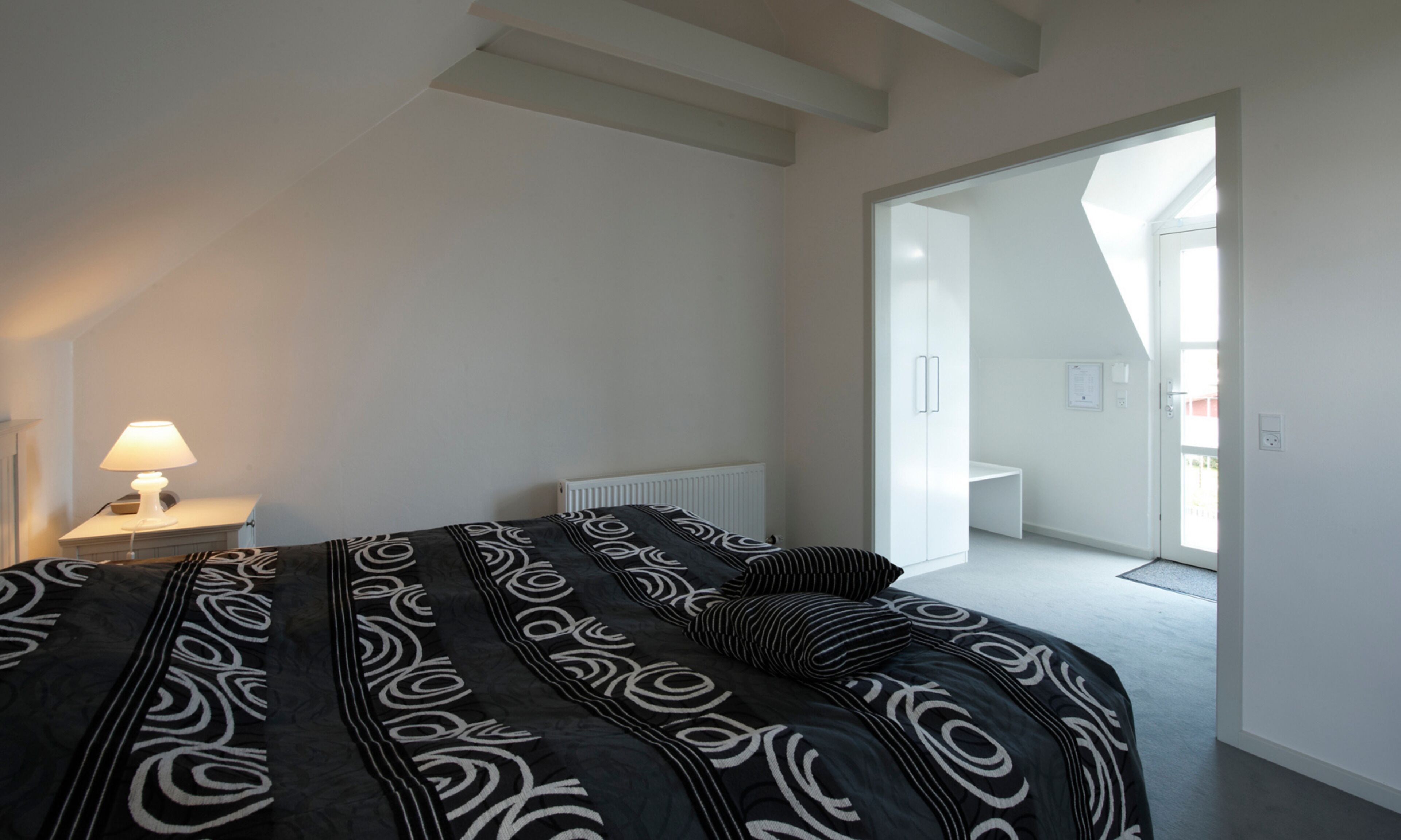 Un dormitorio con una cama, una mesita de noche con una lámpara, un radiador y una puerta que da a otra habitación.