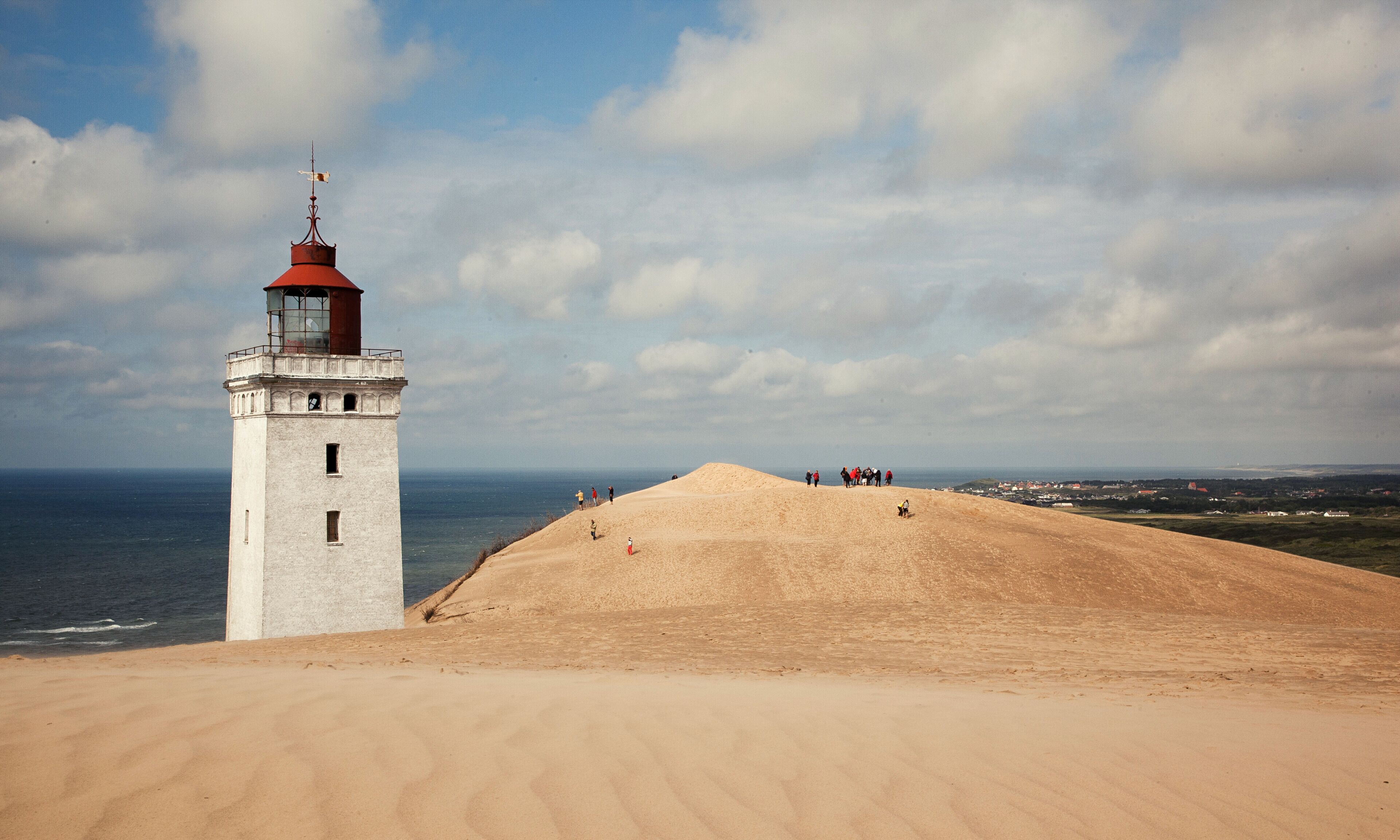Un faro con techo rojo y paredes blancas, ubicado en una colina arenosa con vistas al mar.