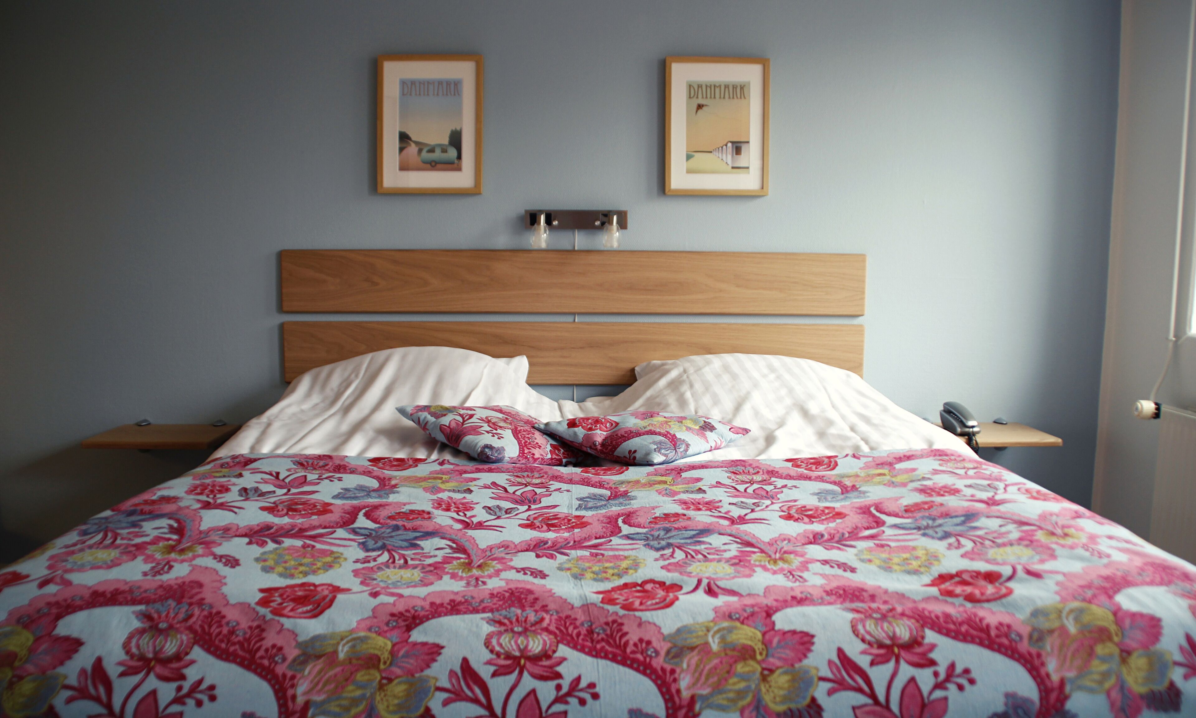 Un dormitorio con cabecera de madera, dos mesitas de noche, colcha con estampado floral y dos cuadros en la pared.