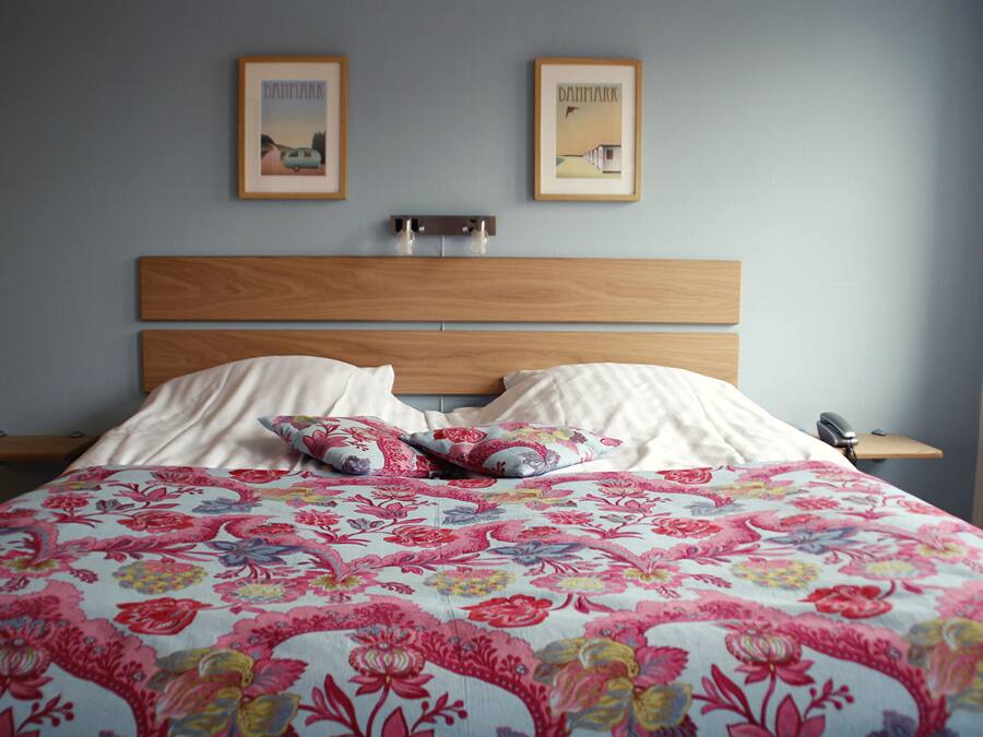 Un dormitorio con cabecera de madera, dos mesitas de noche, colcha con estampado floral y dos cuadros en la pared.