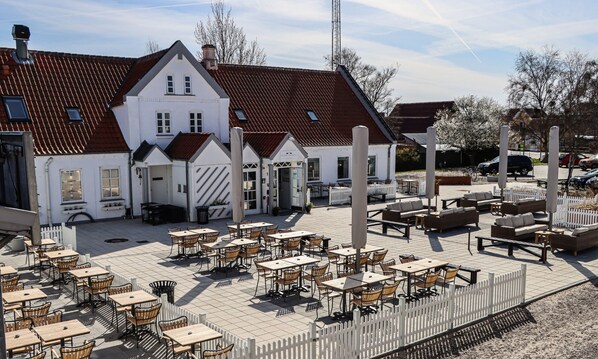 Un edificio de estilo europeo tradicional con tejado de tejas y fachada blanca, con zonas de estar al aire libre con mesas y sillas, y una cerca blanca de piquetes alrededor del patio.