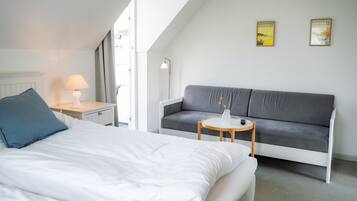 A modern hotel room with a bed, a grey sofa, a small round table, and a lamp. There are two framed pictures on the wall.