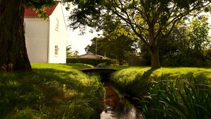 Un jardín tranquilo con un pequeño canal, abundante vegetación y un edificio blanco con techo rojo.