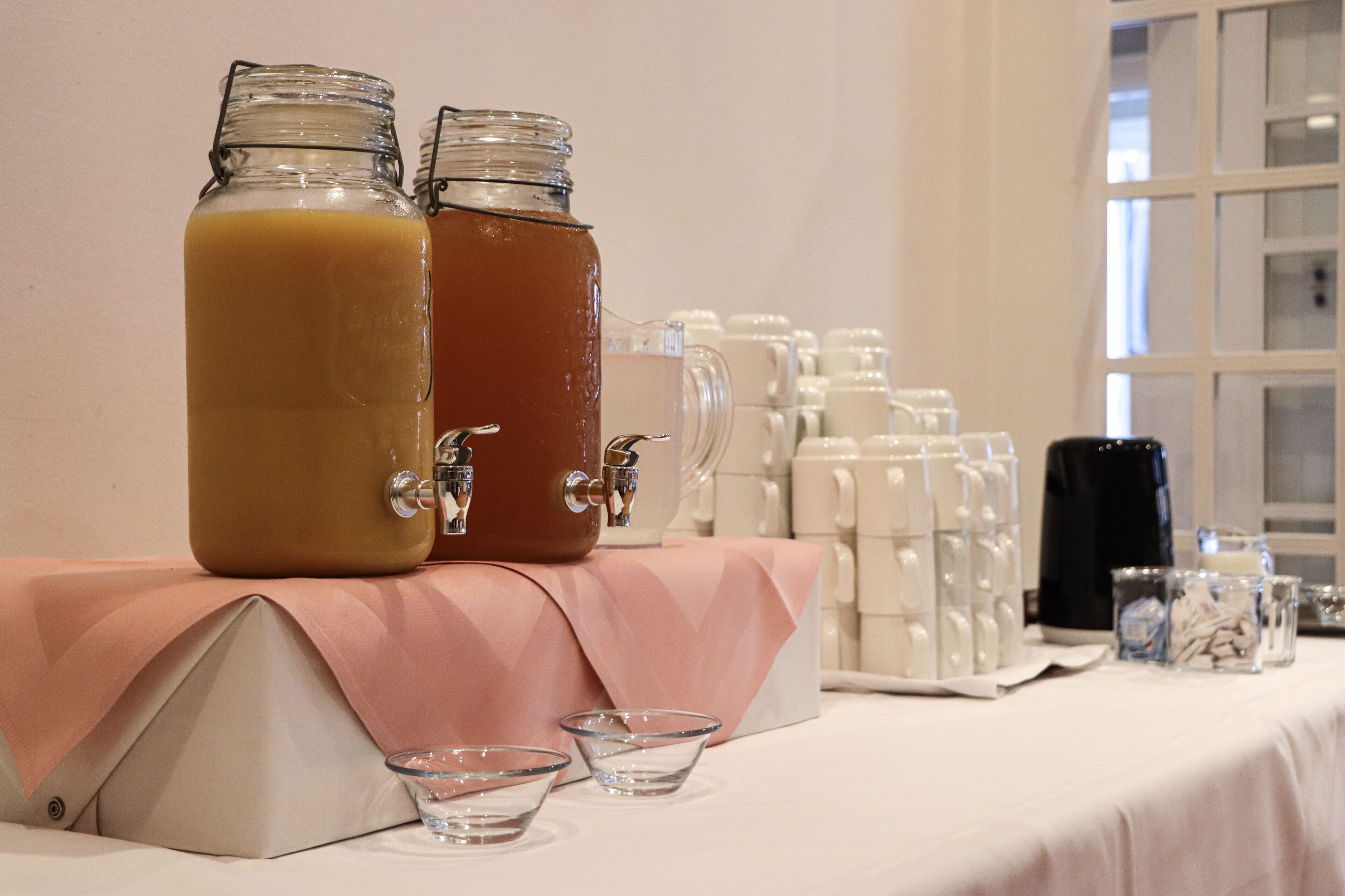 Dos dispensadores de vidrio con grifos metálicos sobre una mesa, rodeados de vasos de vidrio transparentes y un mantel blanco.