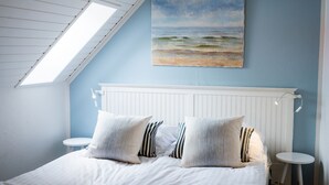 Un dormitorio con techo inclinado, una cama blanca con almohadas a rayas, un cabecero blanco y un cuadro con una escena de playa.