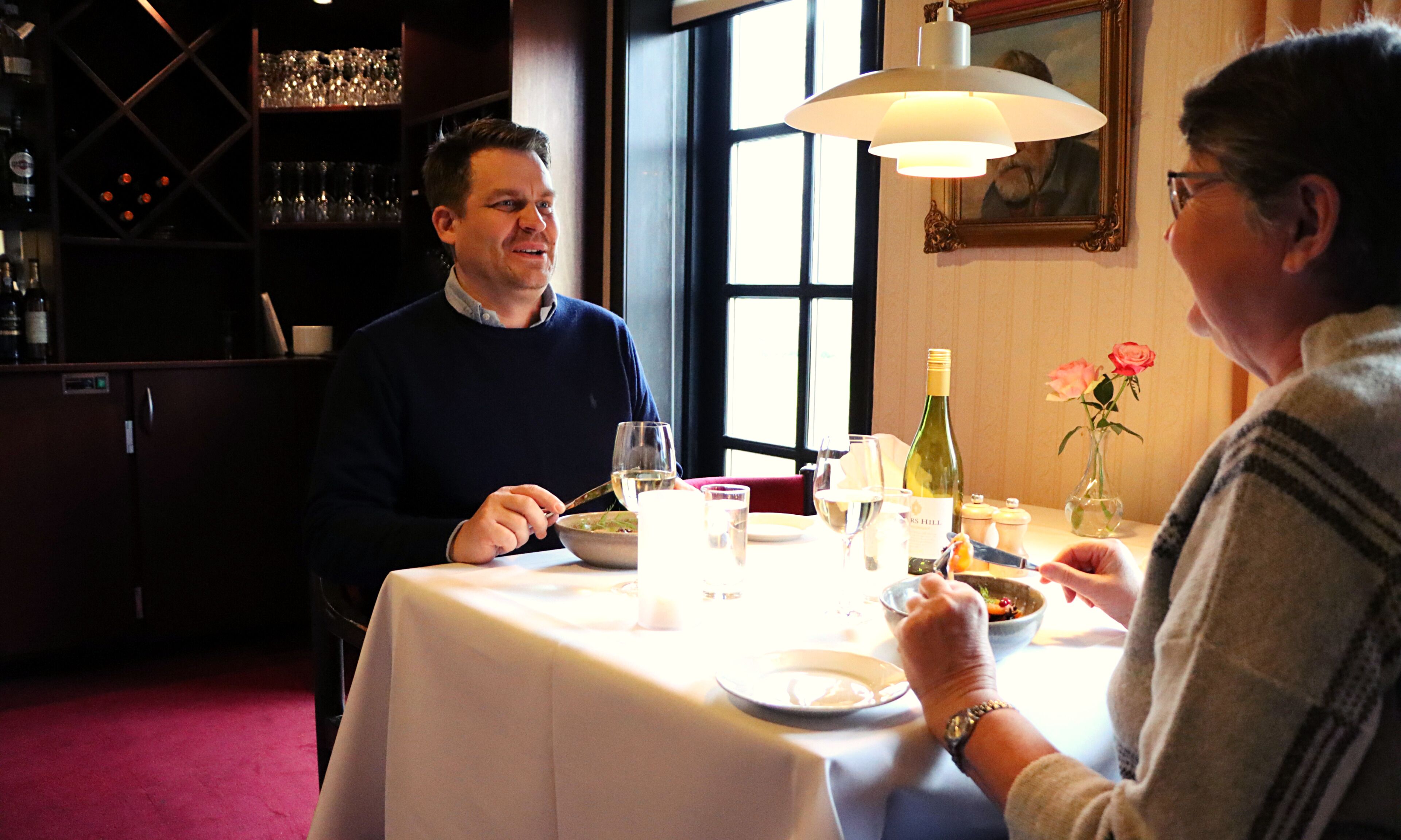 Een eettafel met een witte tafelkleed, wijnflessen, glazen en een vaas met bloemen.
