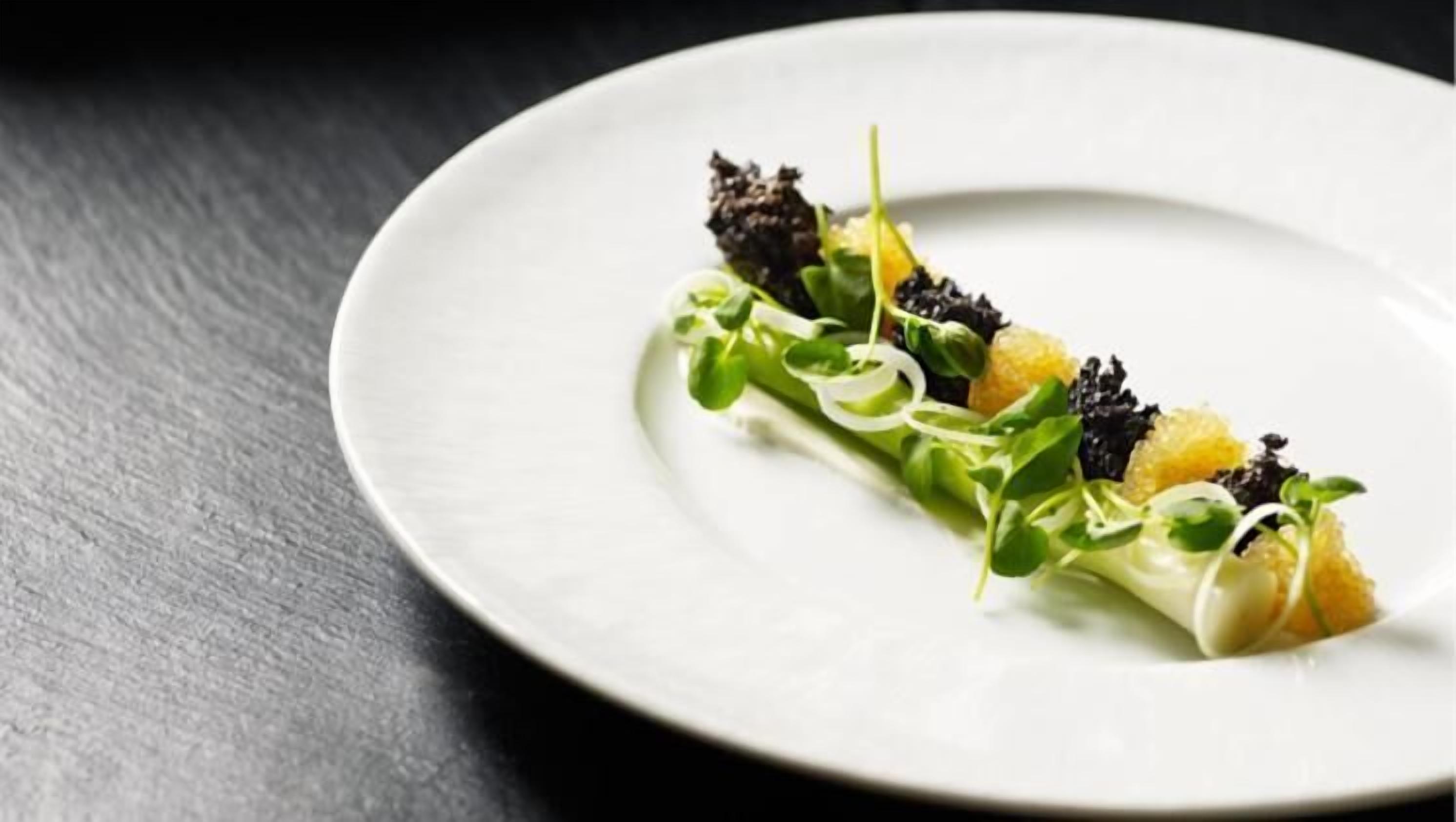 A white plate with a dish of greens and citrus on a dark surface.