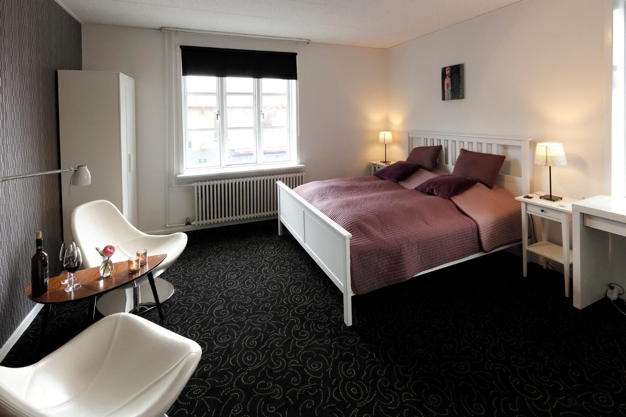 A hotel room with a bed, bedside tables, a desk, a chair, a radiator, and a window.