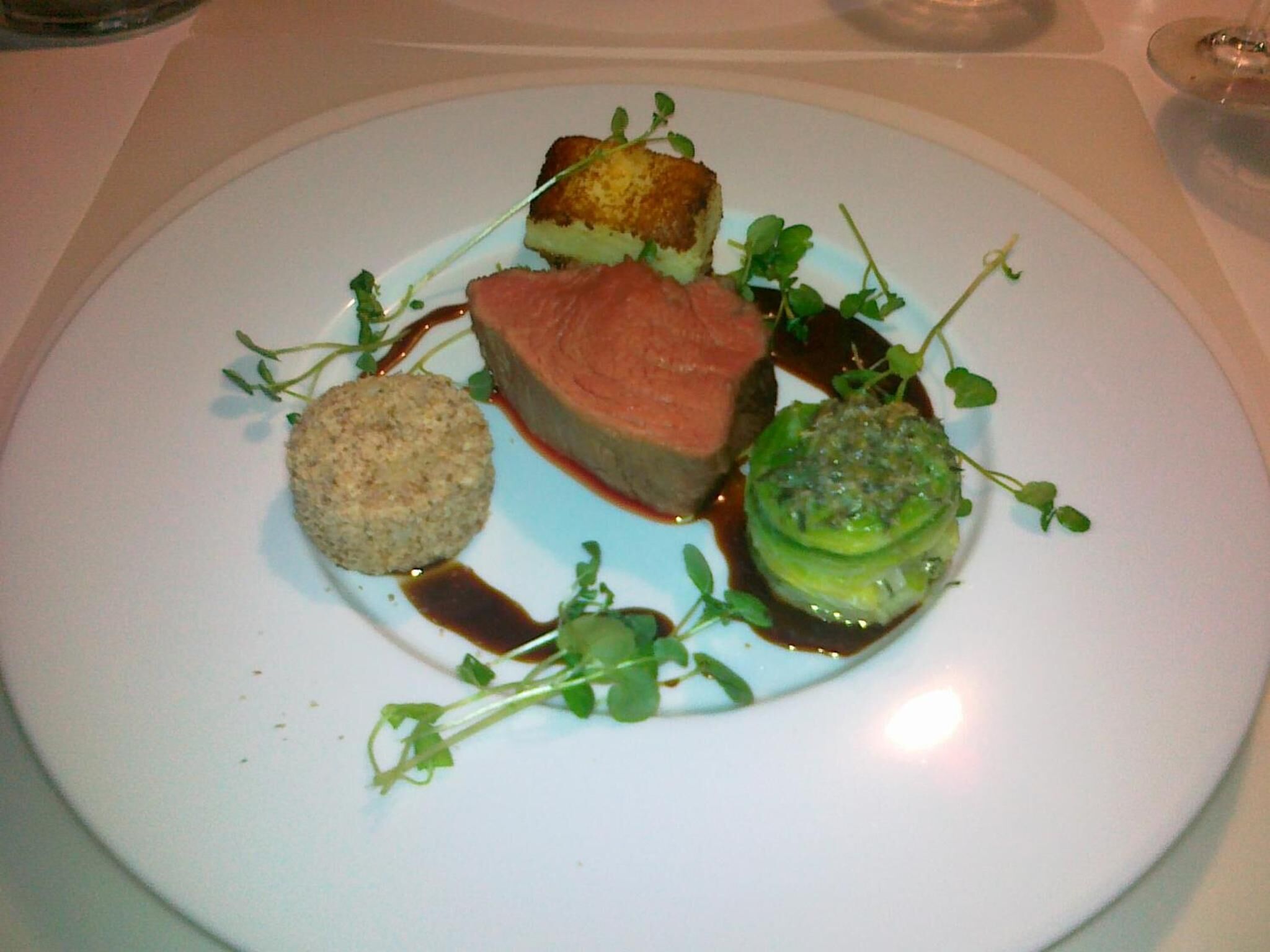 Un plato con un trozo de carne, un elemento redondo y un vegetal verde, todo adornado con hierbas y salsa.