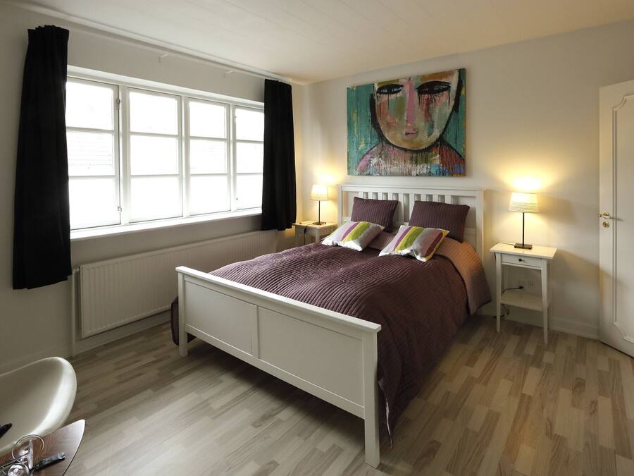 Un dormitorio con cama, dos lámparas de noche, una ventana con cortinas, un radiador y un cuadro en la pared.