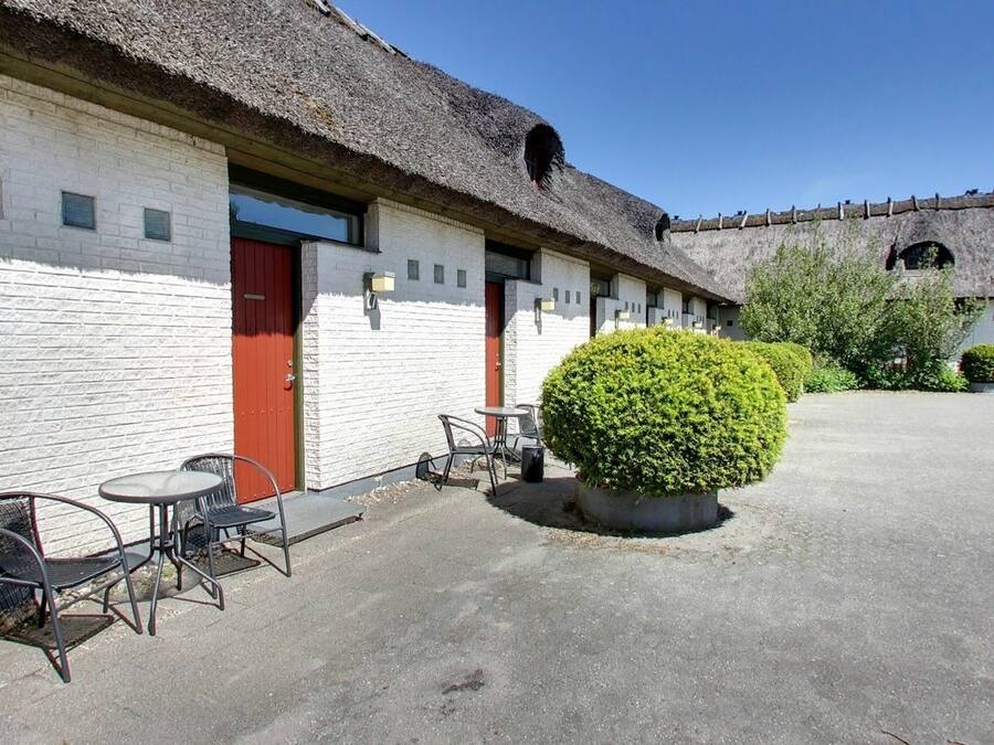 Un edificio con paredes de ladrillo blanco, puertas rojas y techo de paja, con un patio empedrado y un arbusto bien cuidado.