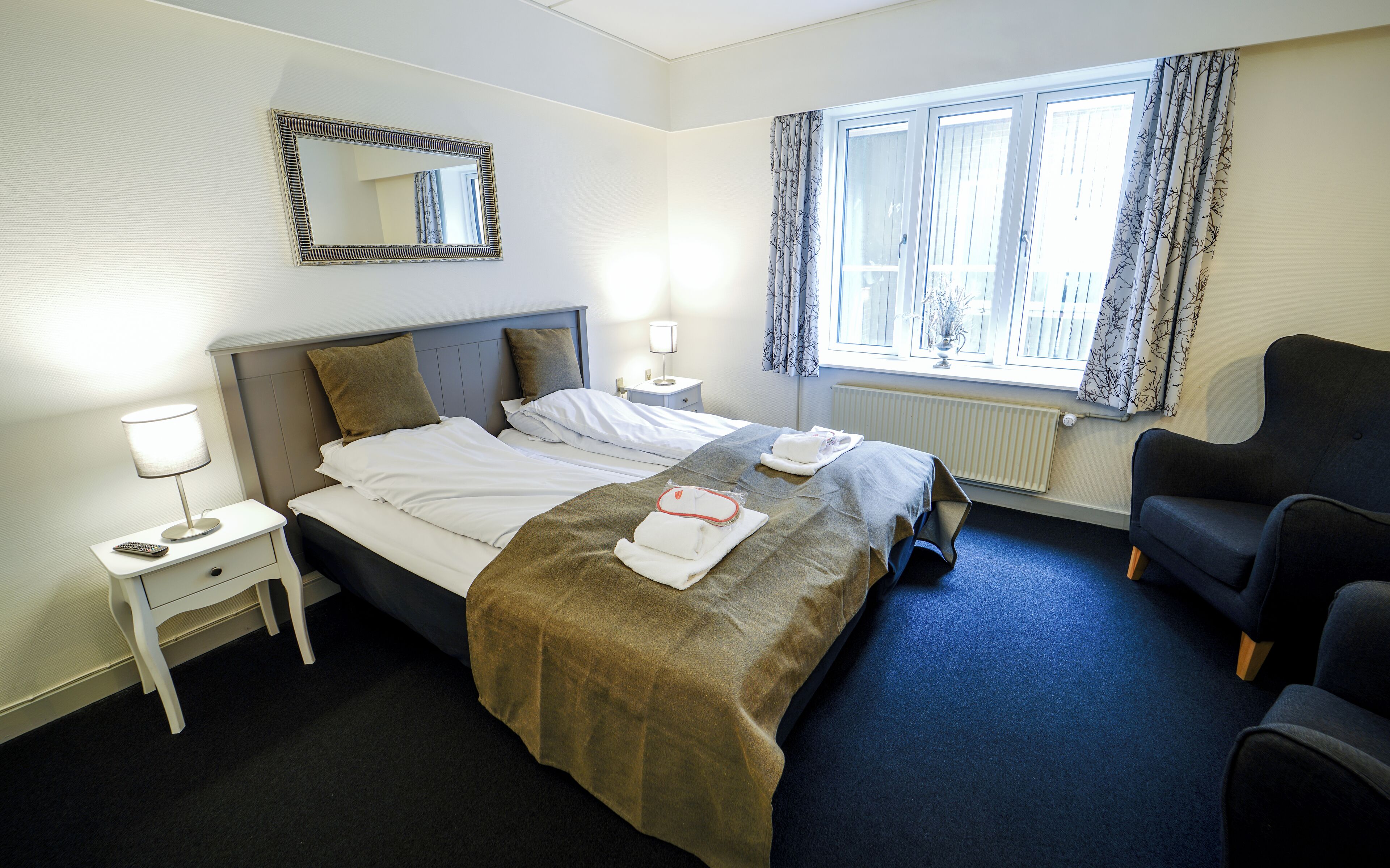 A hotel room with a bed, a chair, a nightstand, a lamp, a mirror, and a window with curtains.