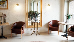 A room with a white brick wall, a large mirror, two leather chairs, a small table with a vase, and a window with a view of a building. - Dronninglund Hotel (Dronninglund)