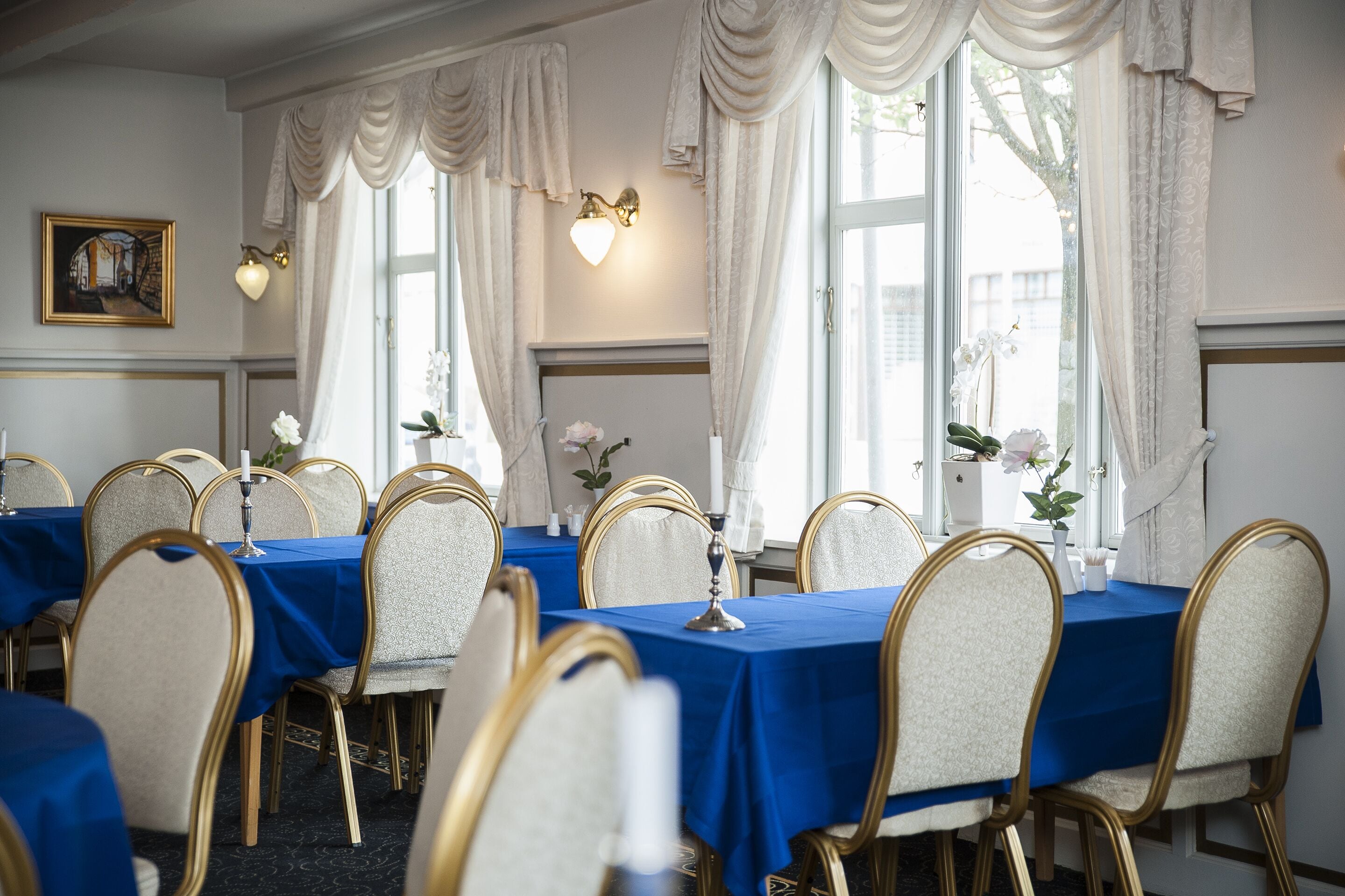 A formal dining room with tables set for six, each with a blue tablecloth and gold chairs.