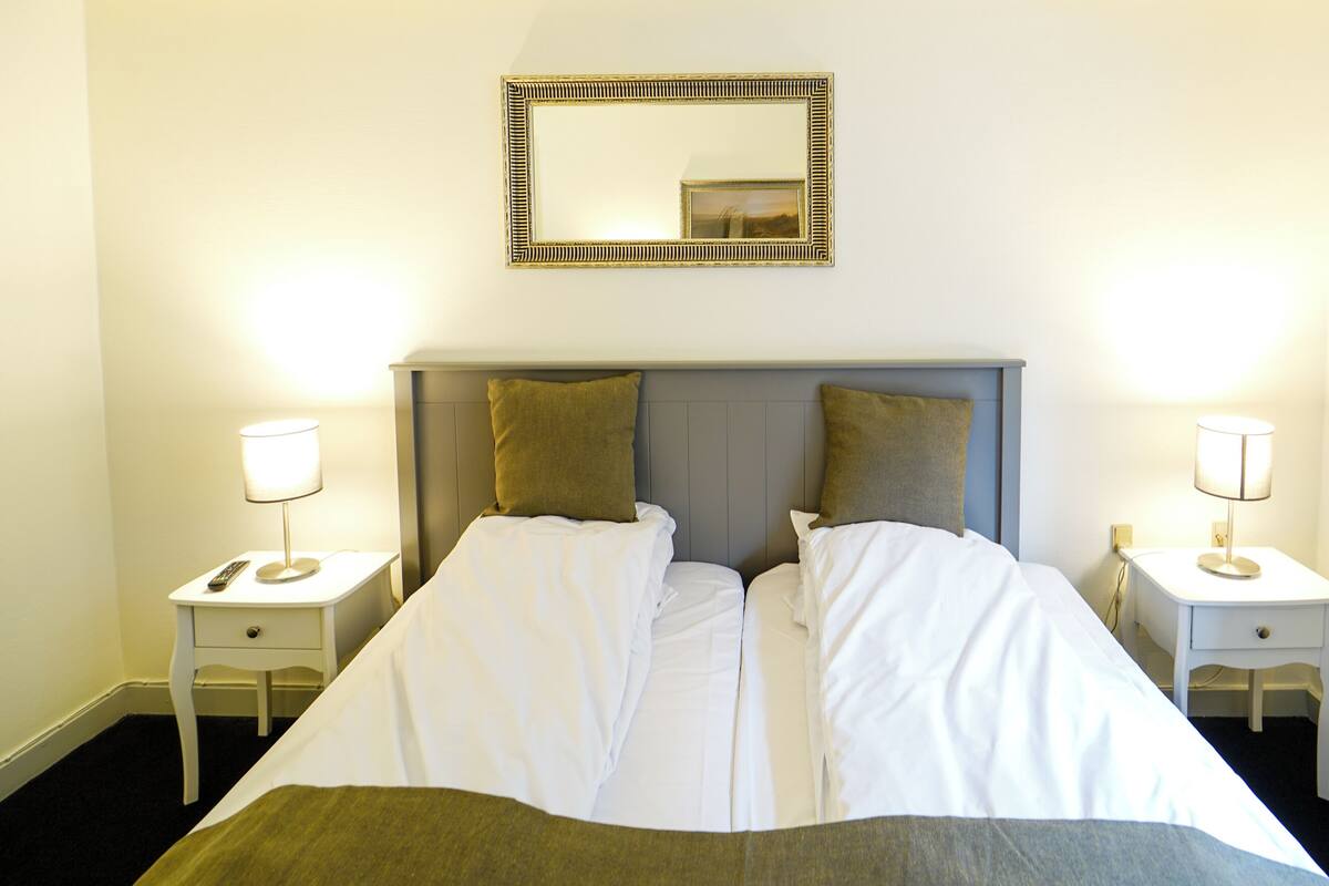 A double bed with white linens, two bedside tables with lamps, a mirror above the bed, and a window with a curtain.
