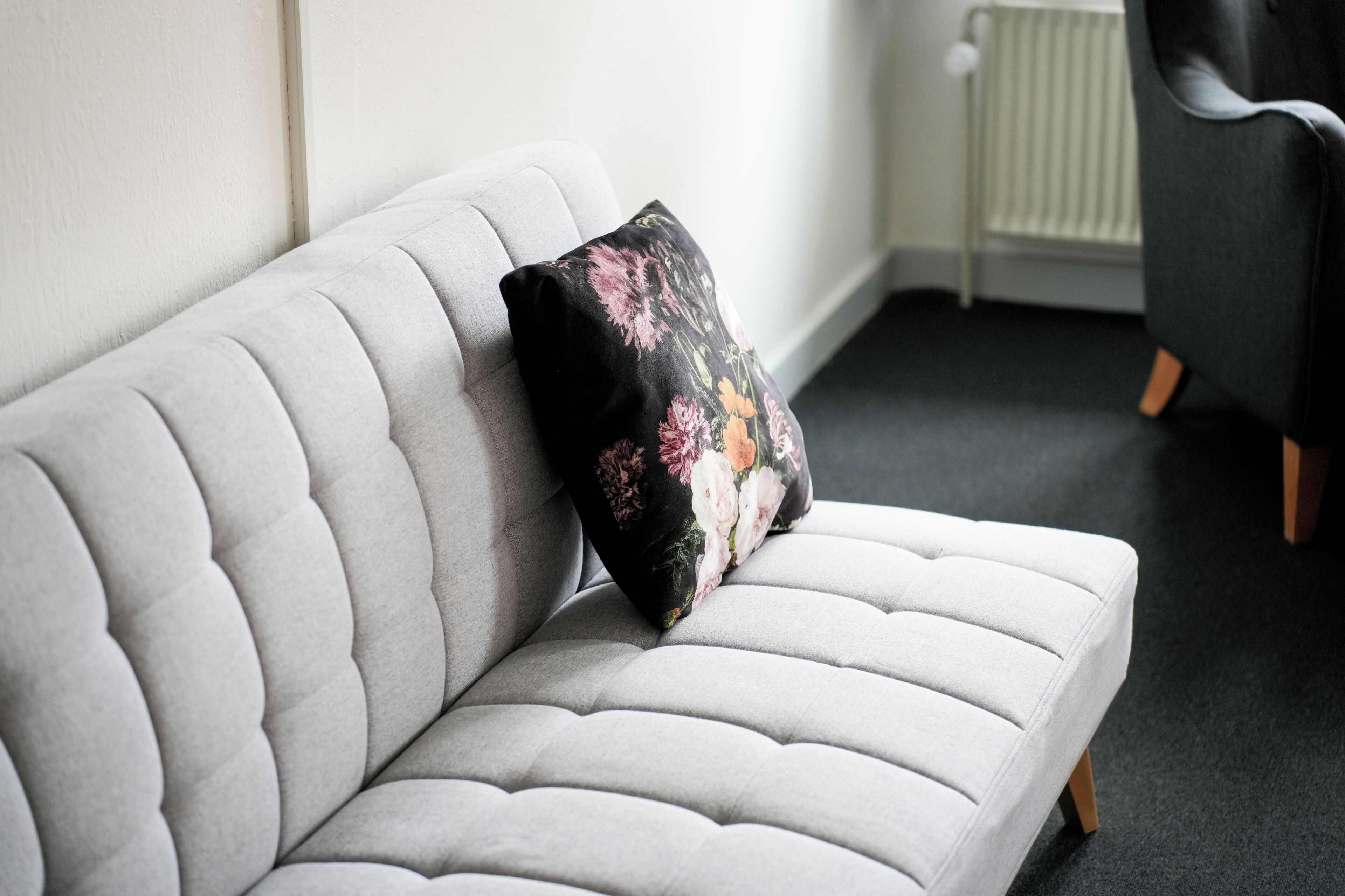 Ein graues, gepolstertes Sofa mit einem floralen Kissen, vor einer weißen Wand mit Heizkörper im Hintergrund.