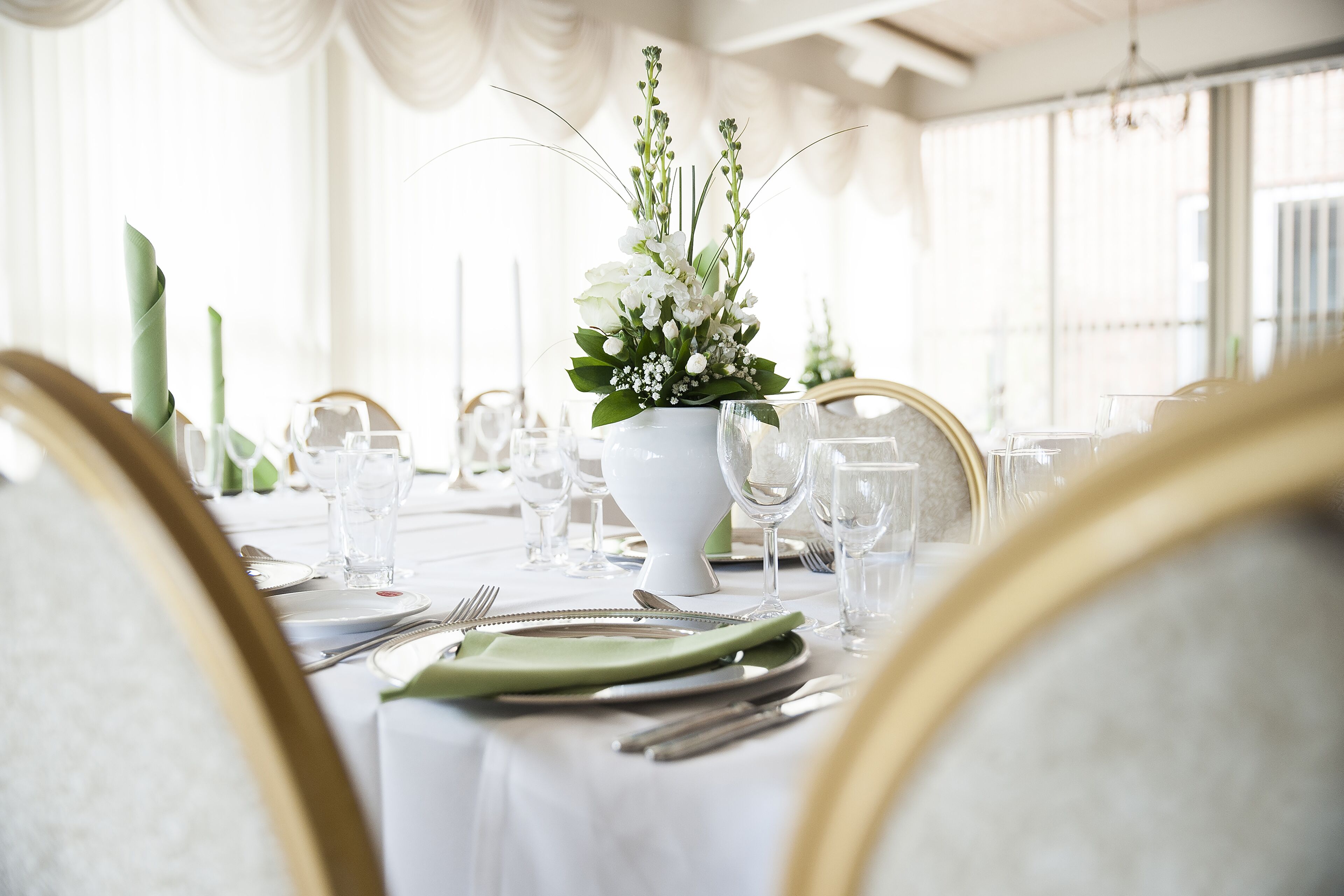 Una mesa de comedor dispuesta con mantel blanco, servilletas verdes y un jarrón blanco con flores.