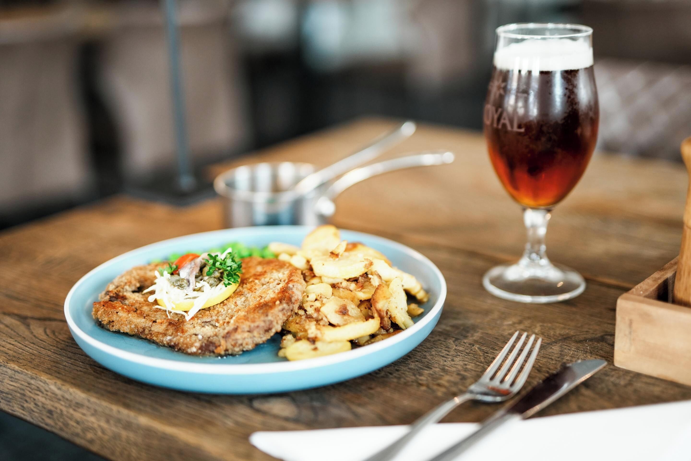 A plate of food with a fried item, potatoes, and vegetables, and a glass of beer on a wooden table.