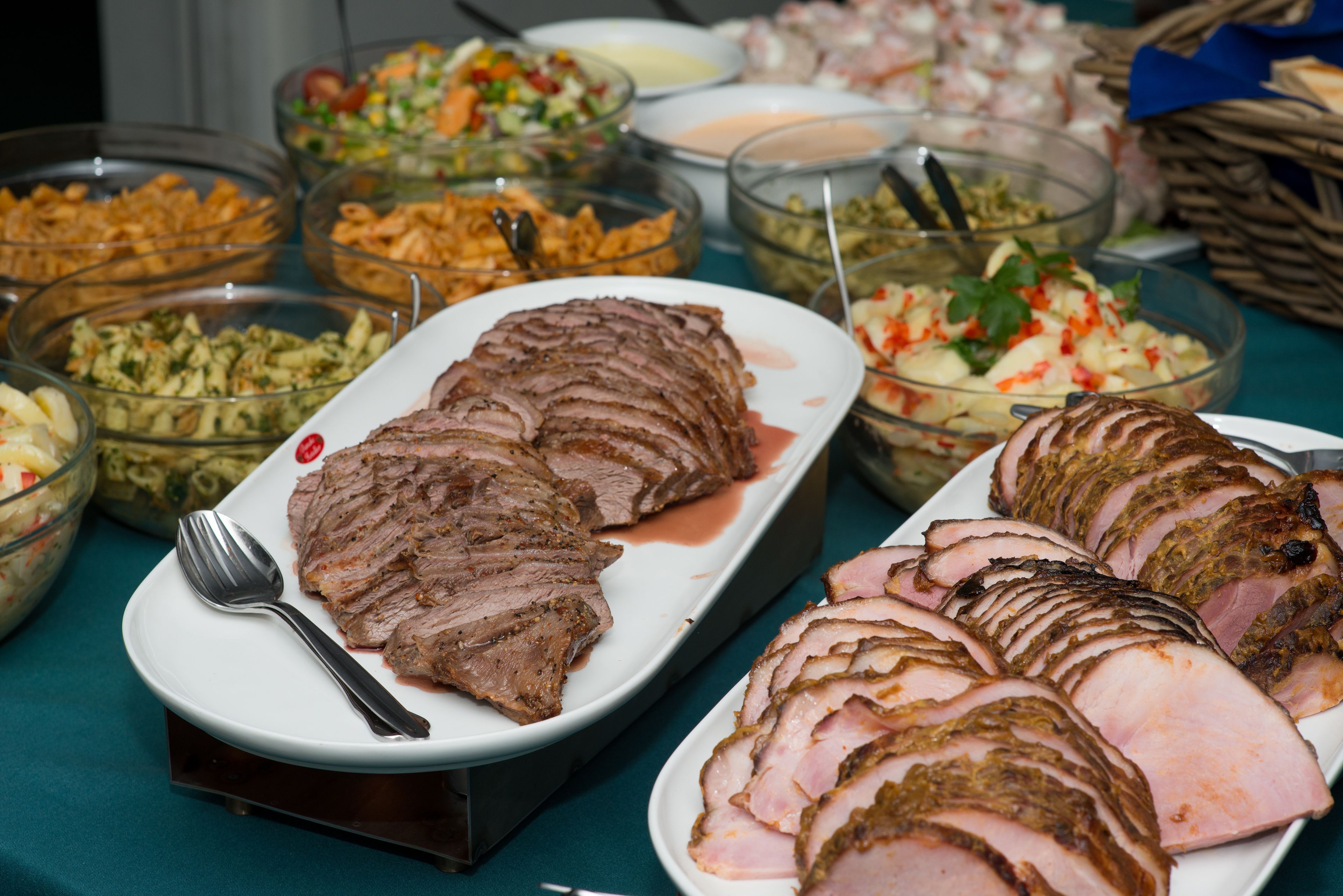 Une table de buffet proposant divers plats, notamment du jambon en tranches, des pâtes et des salades.