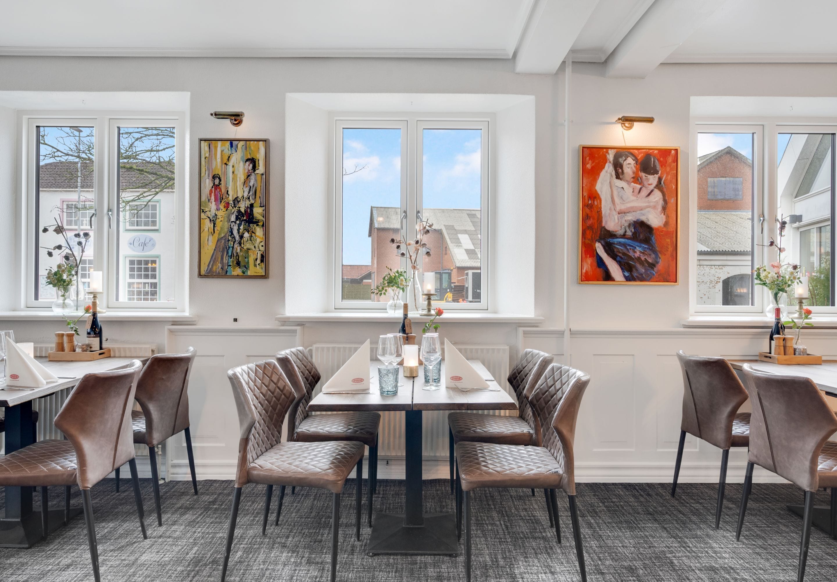 A dining area with a table set for four, surrounded by cushioned chairs, large windows with views of buildings, and artwork on the walls.