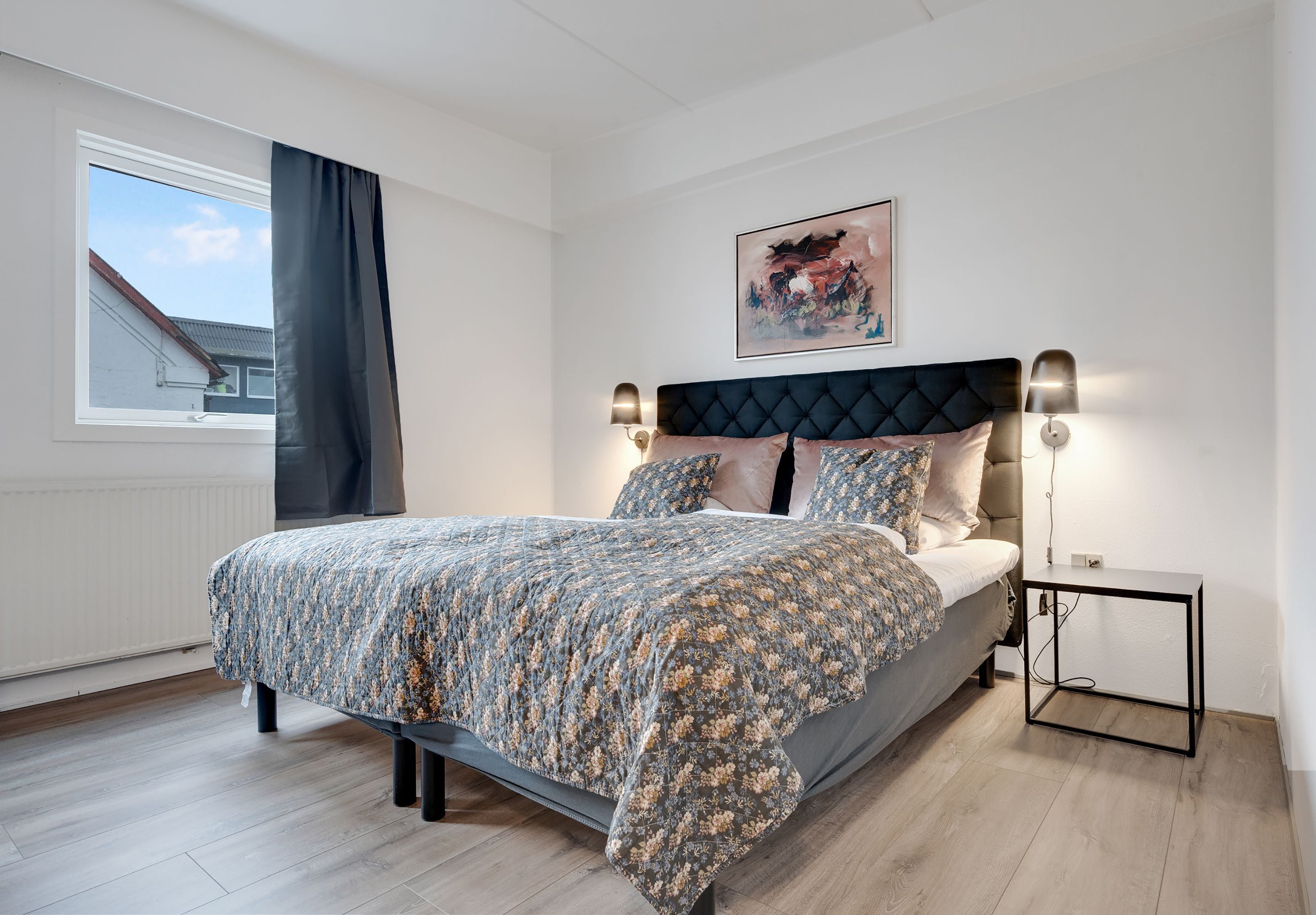 Un dormitorio con cama, mesita de noche, ventana con cortinas, una obra de arte en la pared y un radiador.