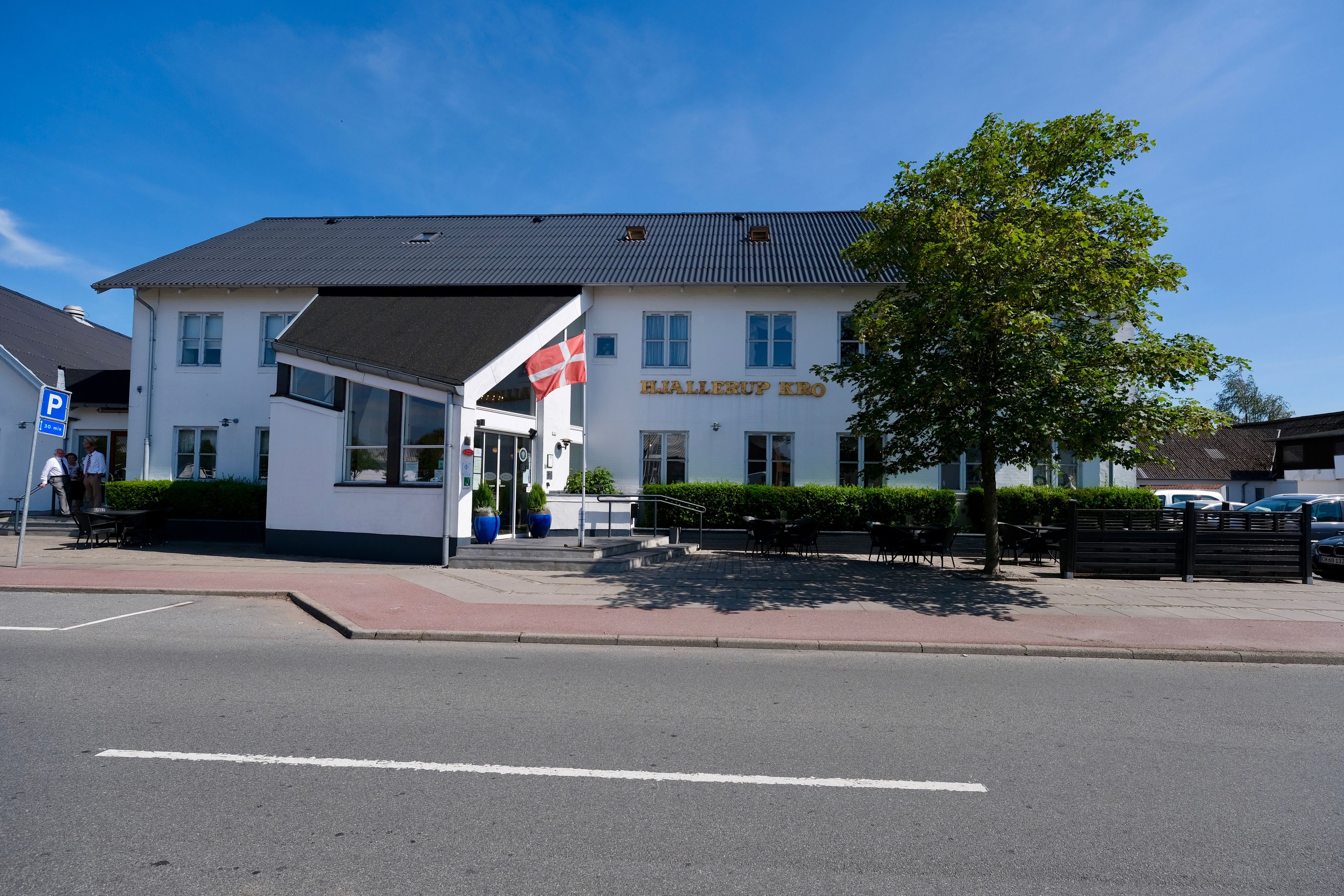 Un edificio con un cartello che riporta la scritta "Hallerup Kro". Ha un tetto piatto, diverse finestre e un marciapiede lastricato di fronte.