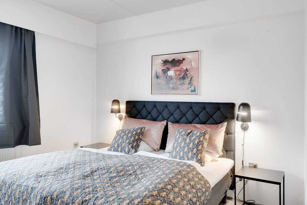 Ein Schlafzimmer mit einer geblümten Bettdecke, einem gepolsterten Kopfteil, zwei Nachttischlampen und einem gerahmten abstrakten Gemälde an der Wand.