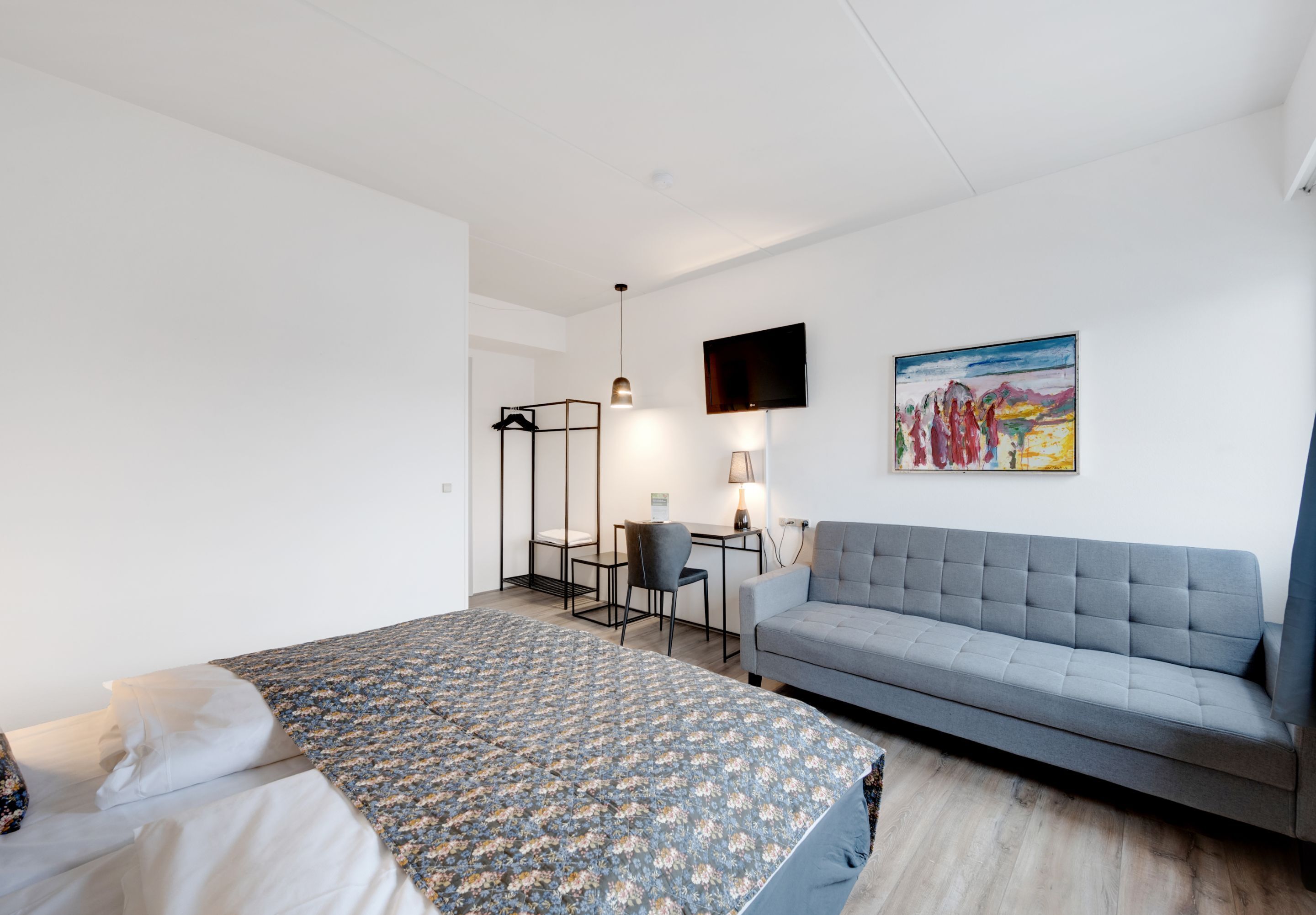 A modern hotel room with a bed, a sofa, a desk with a chair, a TV mounted on the wall, and a painting.