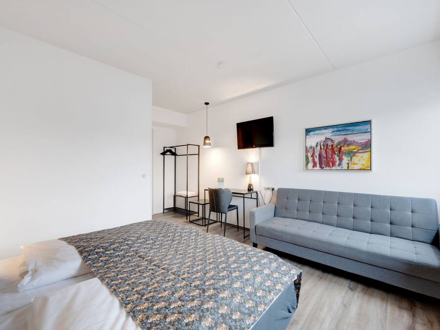 A modern hotel room with a bed, a sofa, a desk with a chair, a TV mounted on the wall, and a painting.