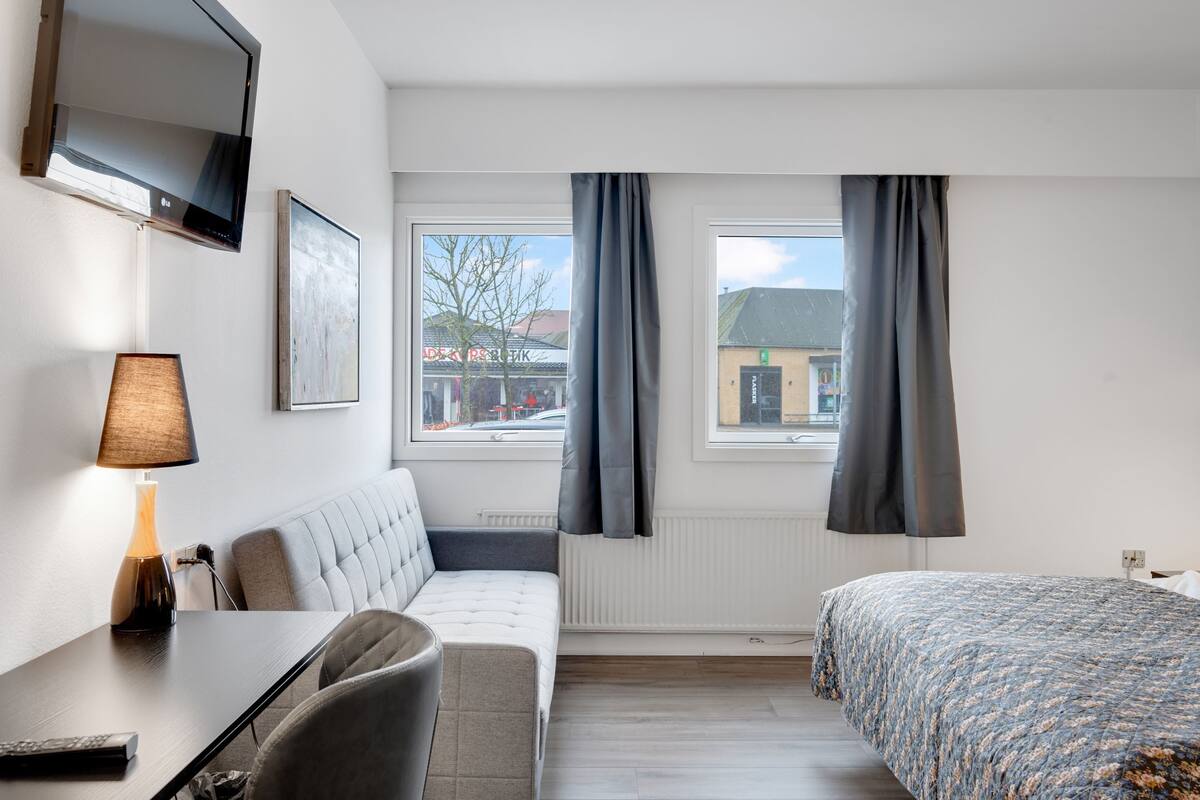 Ein Hotelzimmer mit Bett, einer Sitzecke, Schreibtisch, Fernseher und einem fensterbepflanzten Fenster.