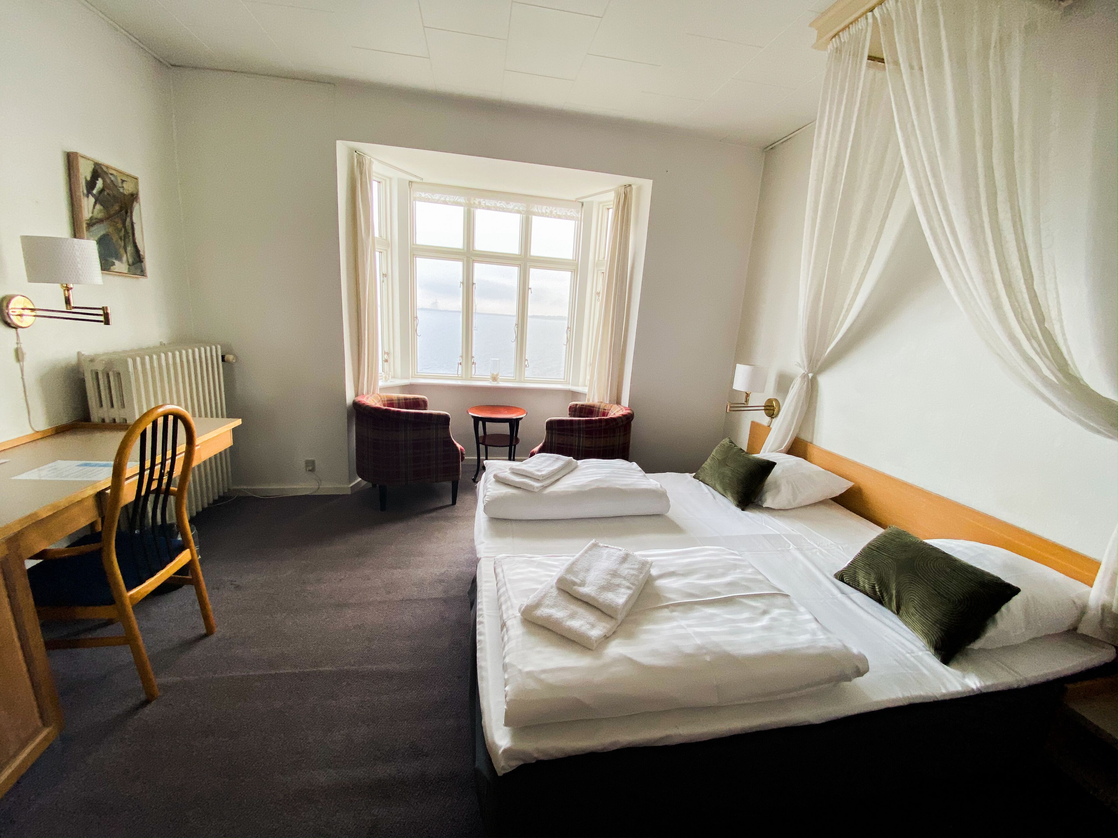 A hotel room with two beds, a desk, a chair, a small table, and a view of the sea through a large window.