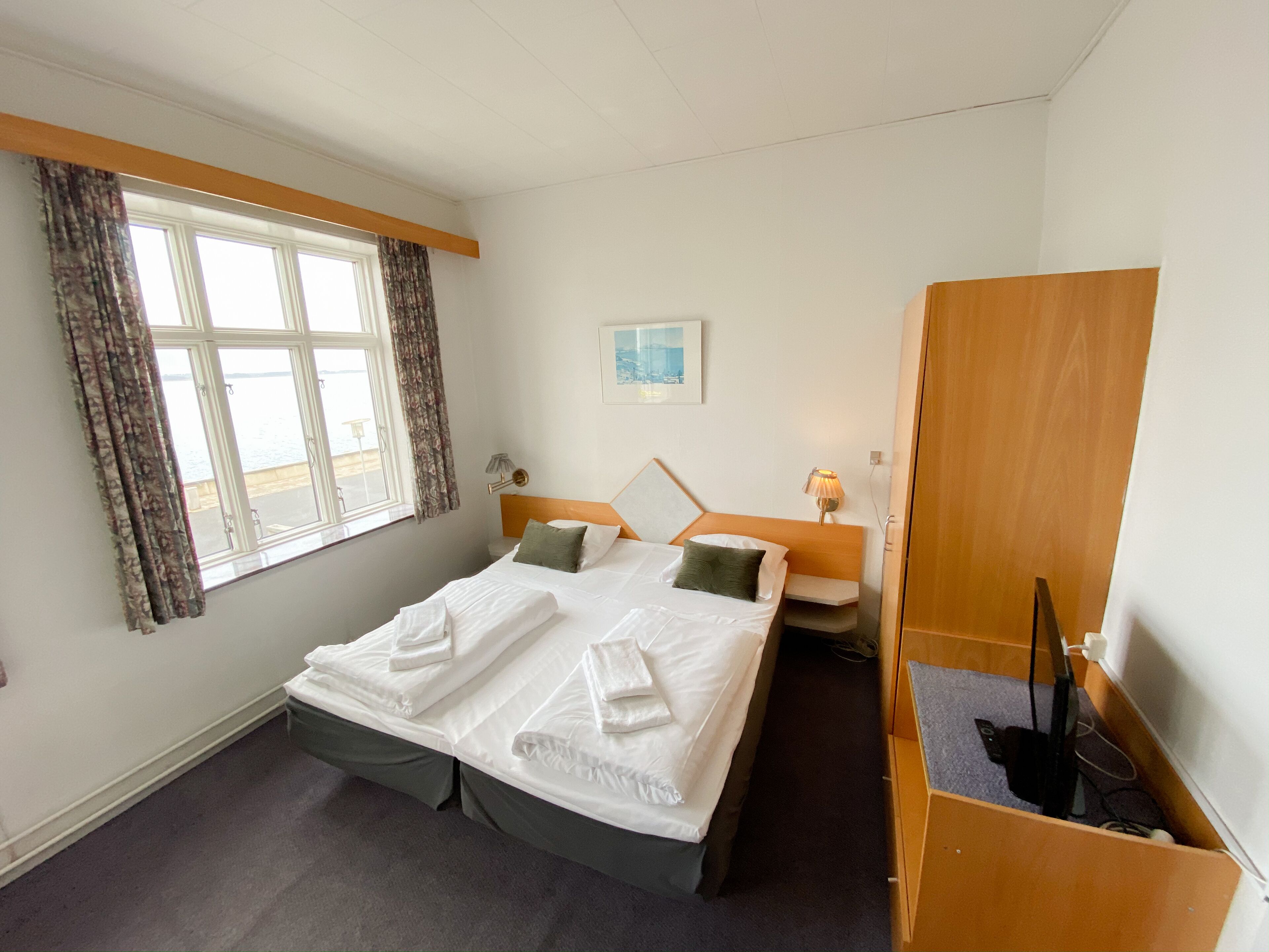 Ein Hotelzimmer mit Bett, Schreibtisch, Stuhl, einem Fenster mit Vorhängen und einem Bild an der Wand.