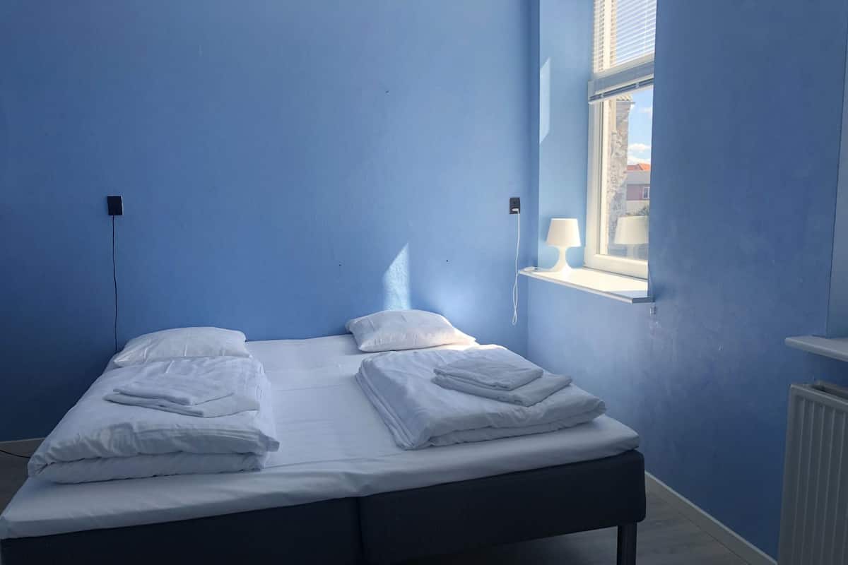 Una cama individual con sábanas blancas, una mesita de noche con lámpara y una ventana con persianas.