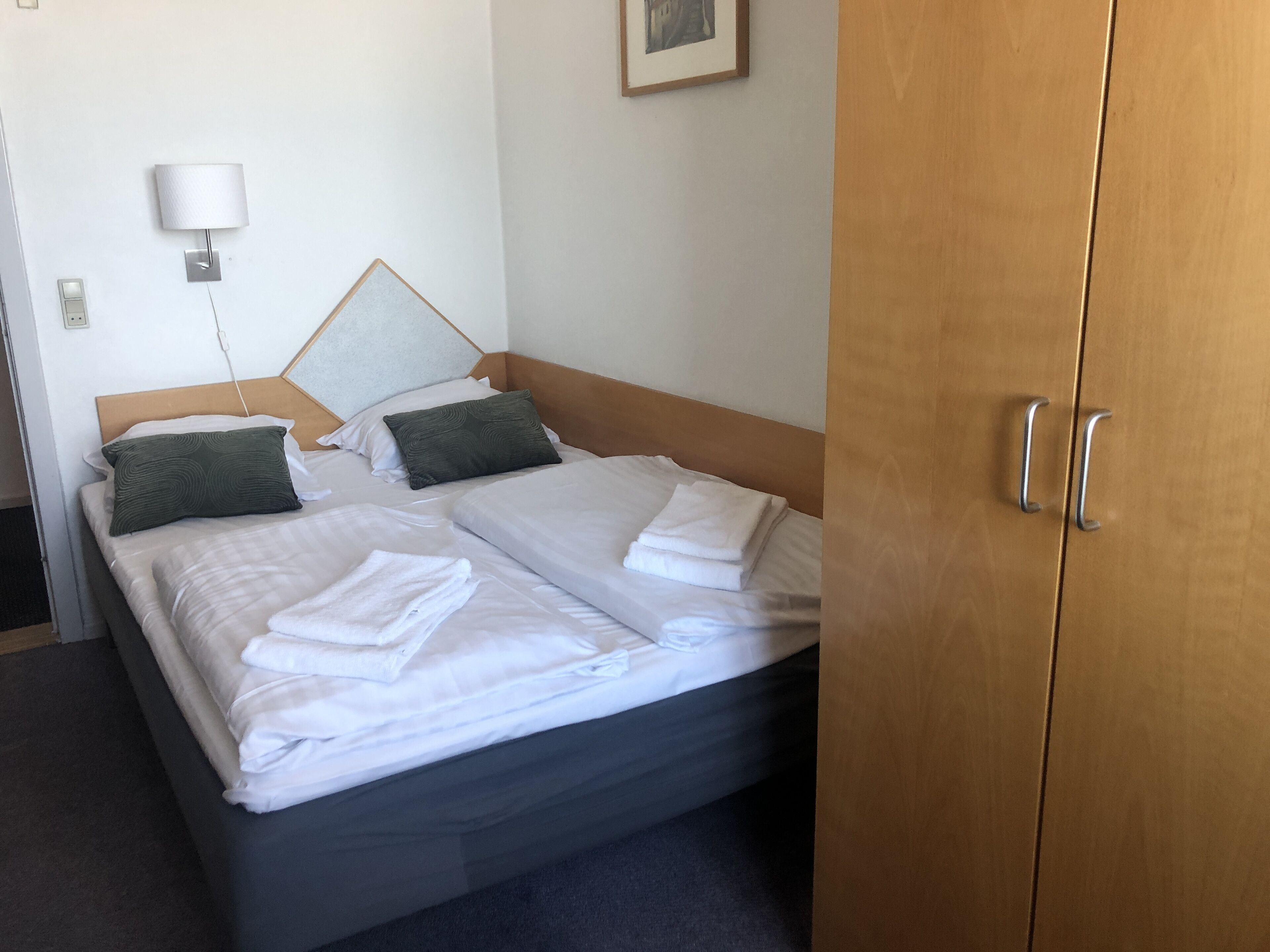 Ein ordentlich bezogenes Bett mit weißen Leinen und zwei Kissen, ein hölzernes Kopfteil, eine Nachttischlampe und ein zweitüriger Holzschrank.
