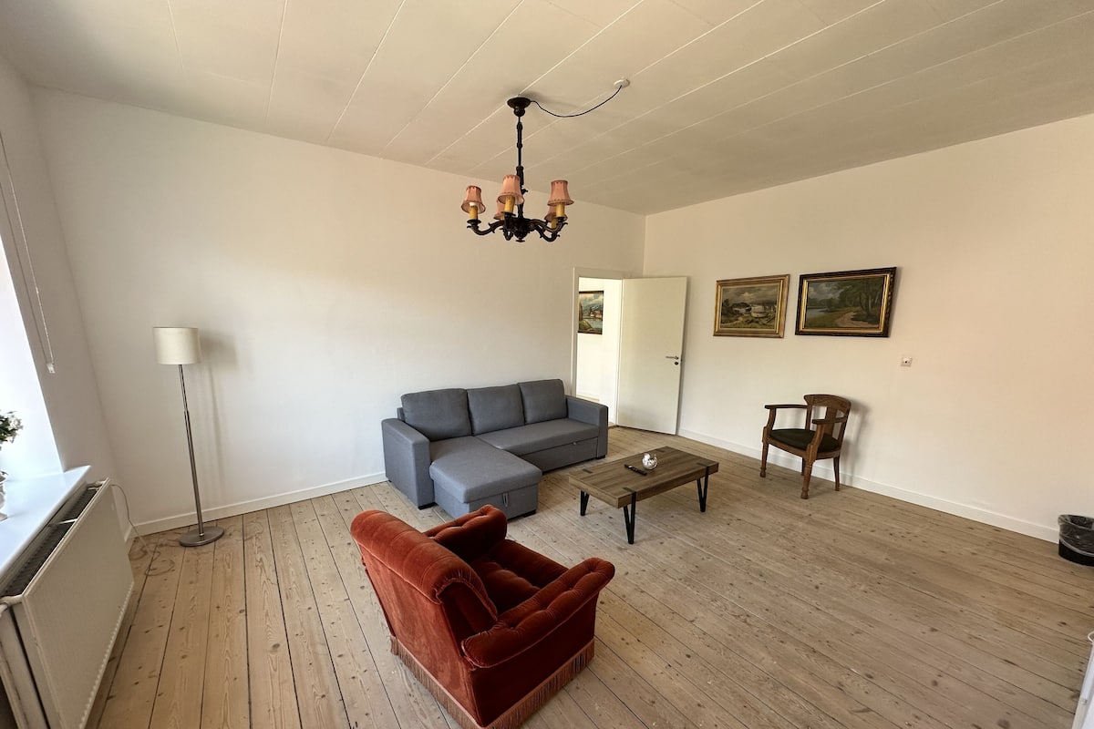 Una sala de estar con un sofá seccional gris, un sillón de terciopelo rojo, una mesa de centro de madera, una lámpara de pie y dos cuadros en la pared.
