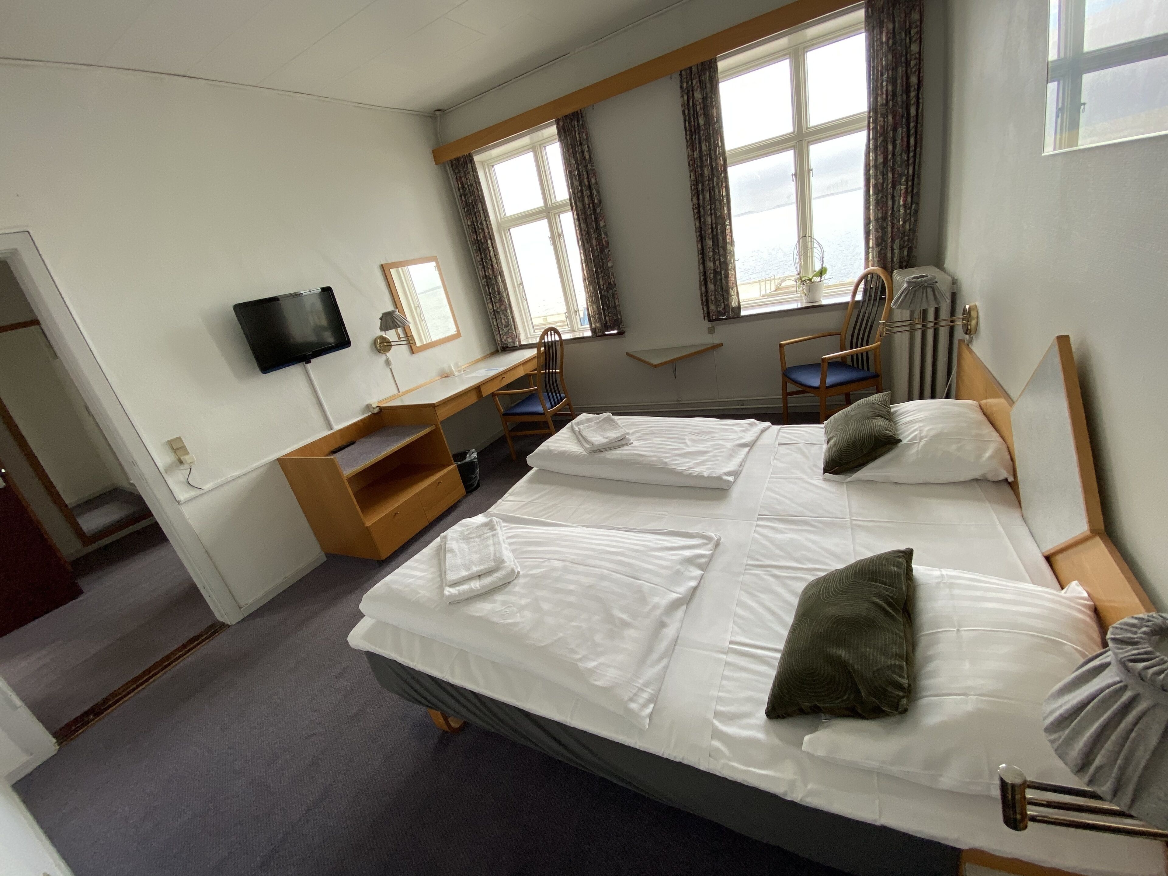 Ein Hotelzimmer mit zwei Betten, einem Schreibtisch, einem Stuhl, einem Fernseher und großen Fenstern mit Vorhängen.