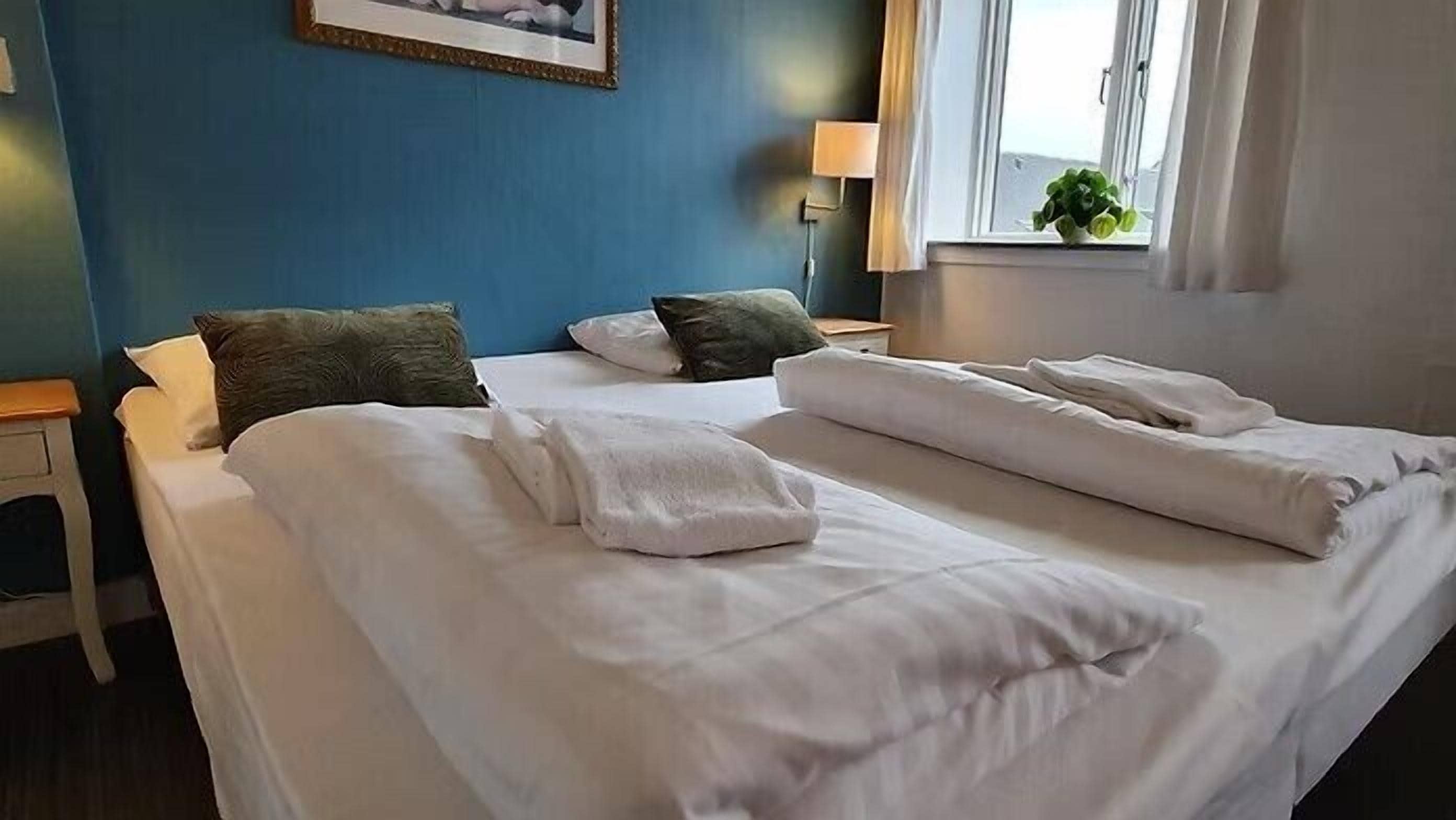 Camera d'albergo con due letti, un comodino, una lampada e una finestra con una pianta.