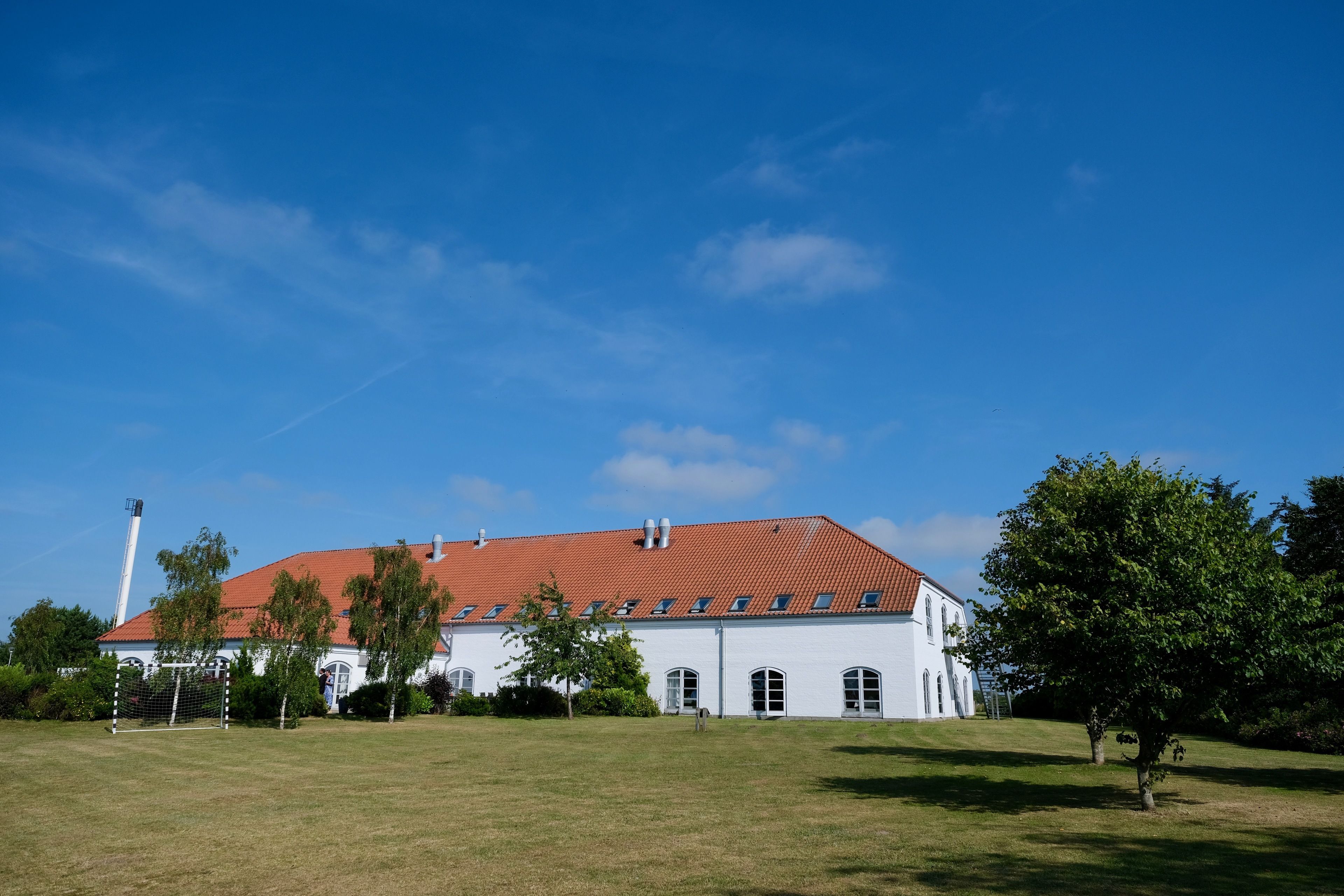 Un grand bâtiment blanc, surmonté d’un toit rouge, entouré d’un champ verdoyant et d’arbres.