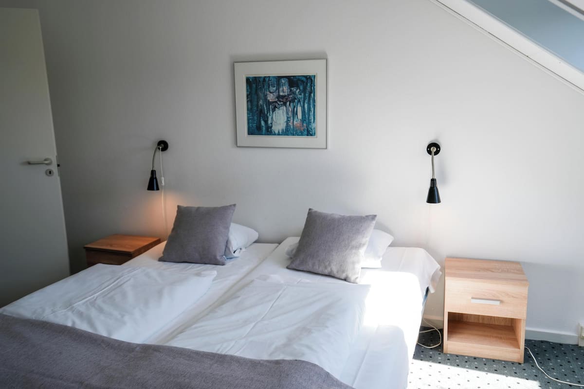Un dormitorio con cama, dos mesitas de noche, lámpara de pared y un cuadro.