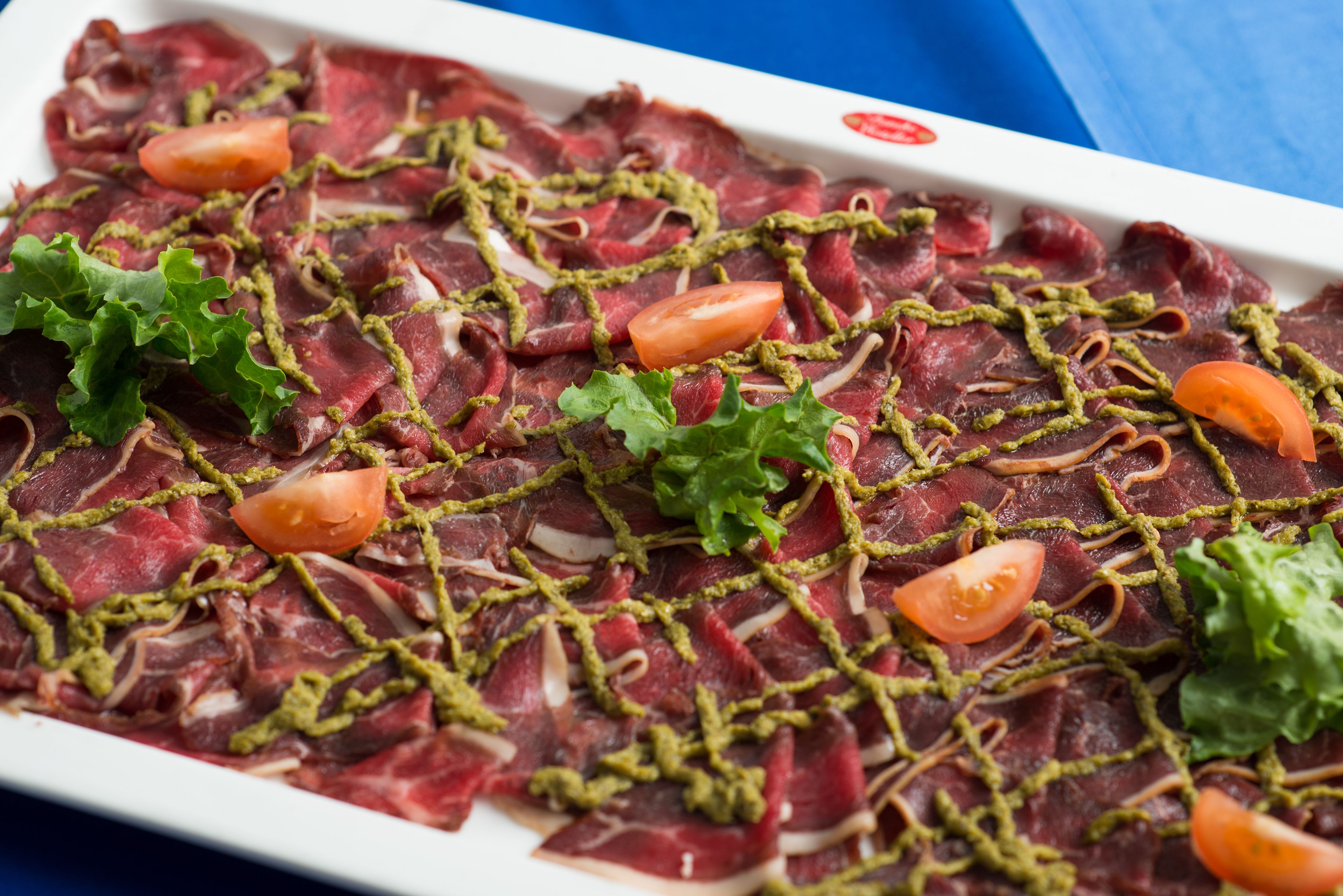 Un plato rectangular blanco con carne roja en lonjas finas, adornado con hierbas verdes y rodajas de tomate.