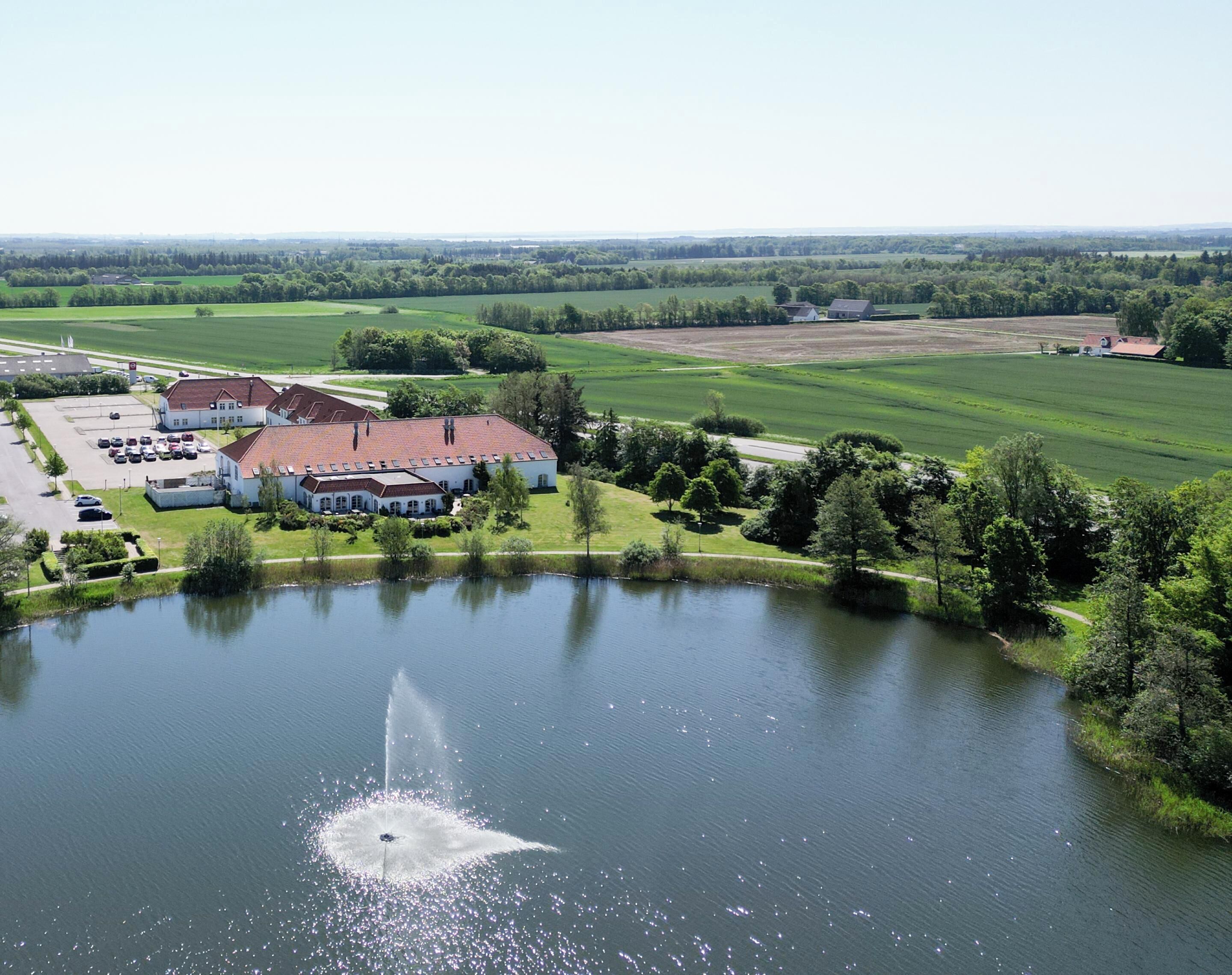 Eine Luftaufnahme einer Anlage mit einem großen Gebäude, einem Teich mit einer Springbrunnen und umliegenden grünen Feldern.