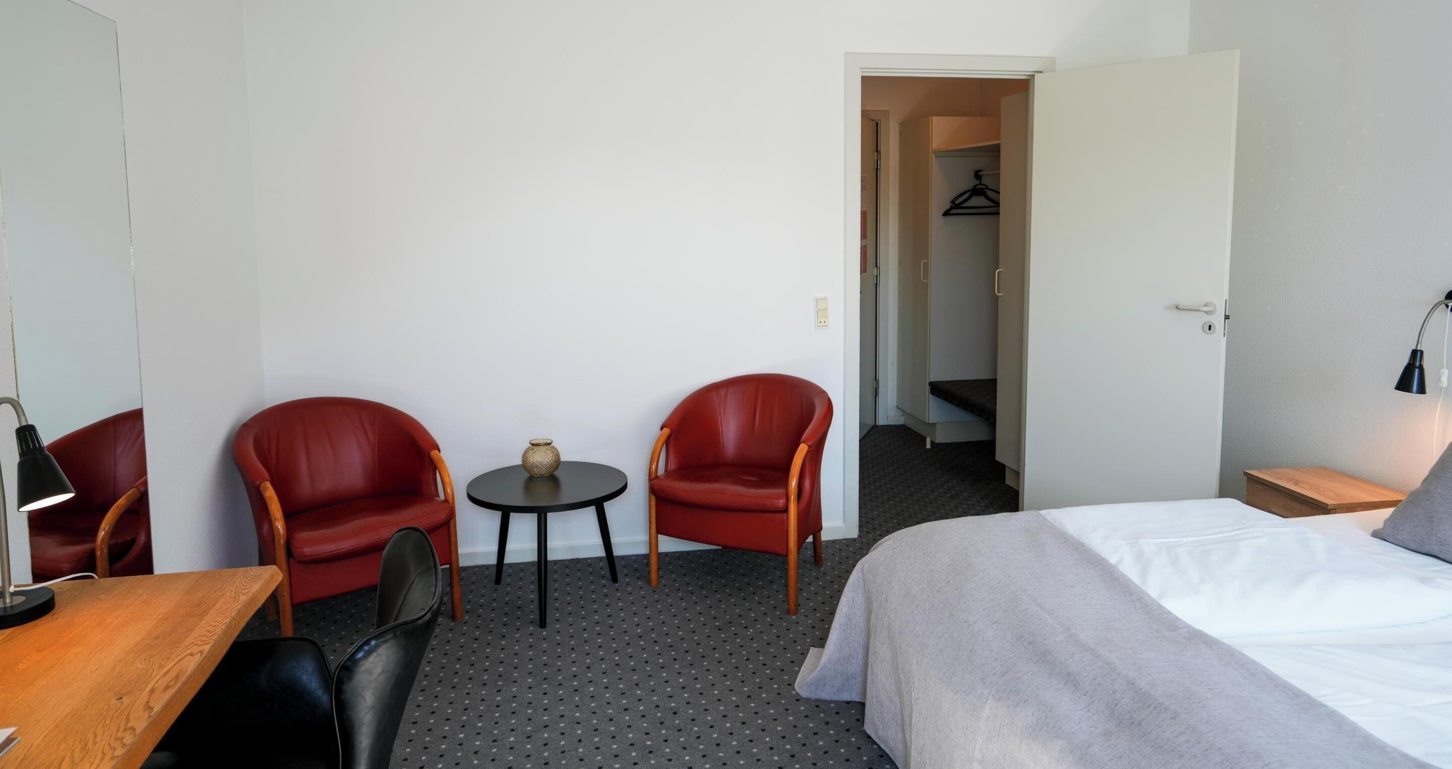 Ein Hotelzimmer mit einem Bett, zwei roten Sesseln, einem kleinen Tisch mit einer Vase, einem Schreibtisch mit einer Lampe und einem Schrank mit einem Kleiderbügel.