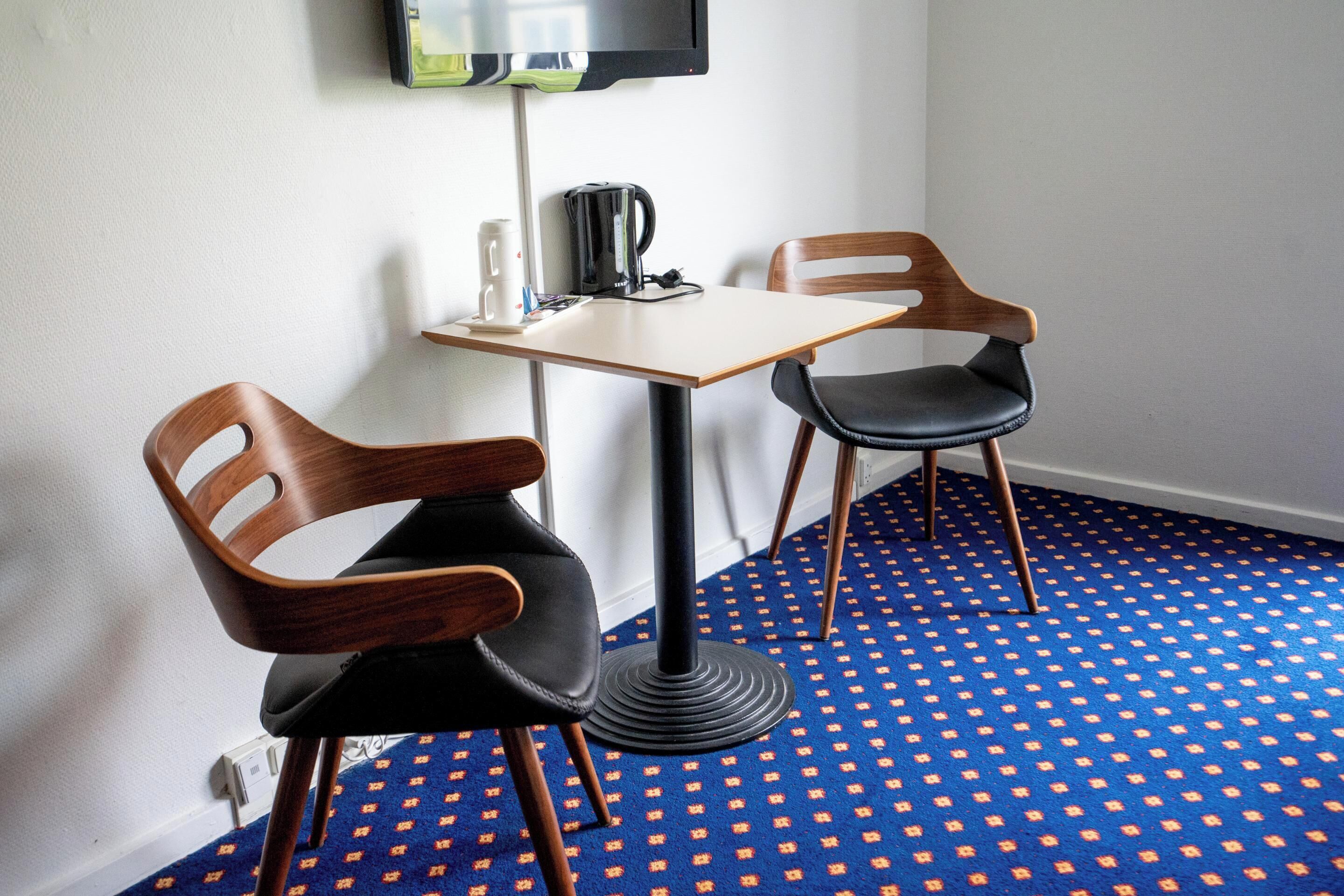 A room with a small table and two chairs, a television mounted on the wall, and a patterned carpet.