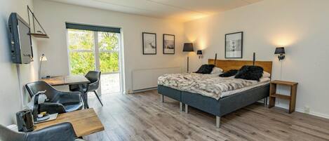 A modern bedroom with a bed, a desk, and a chair. There are framed pictures on the wall and a window with a view of greenery.