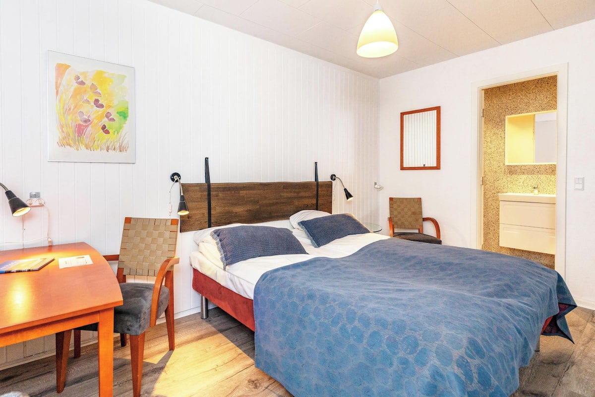 A bedroom with a bed, a desk, a chair, a lamp, a mirror, and a bathroom with a sink and mirror.