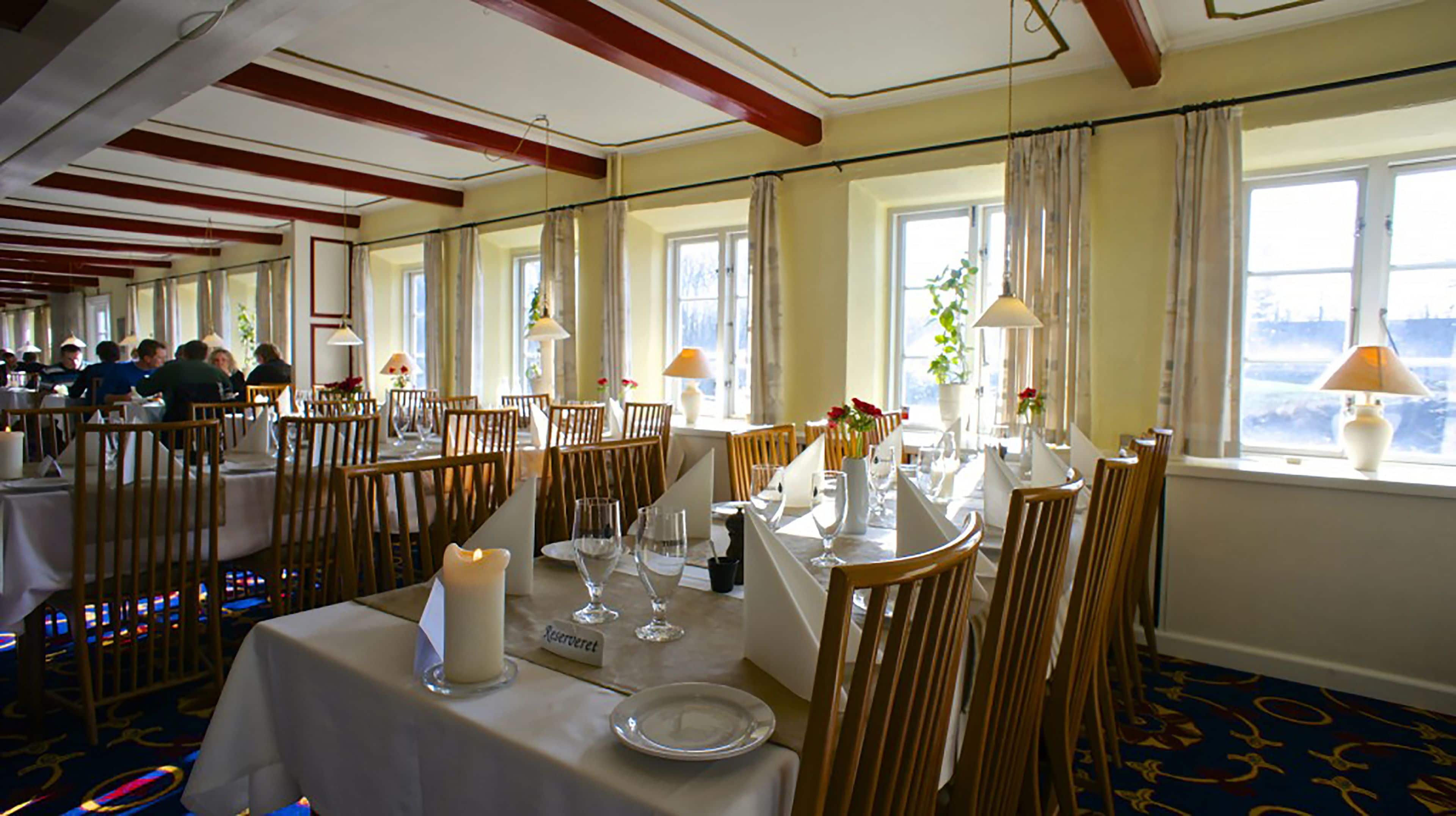 A dining area with tables set for service, wooden chairs, and a view of the outdoors through large windows.