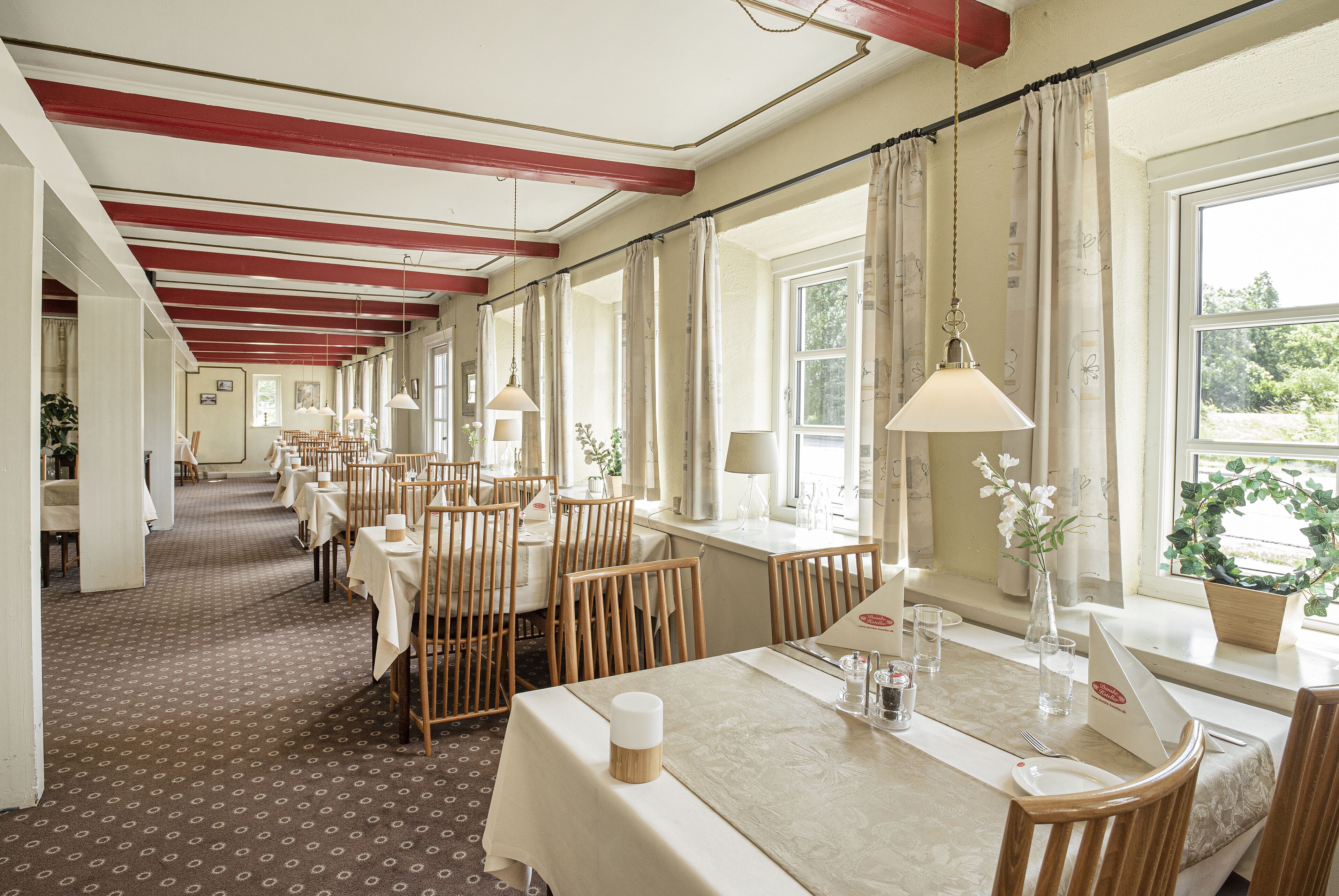 A hotel dining area with long tables, wooden chairs, and hanging lamps.