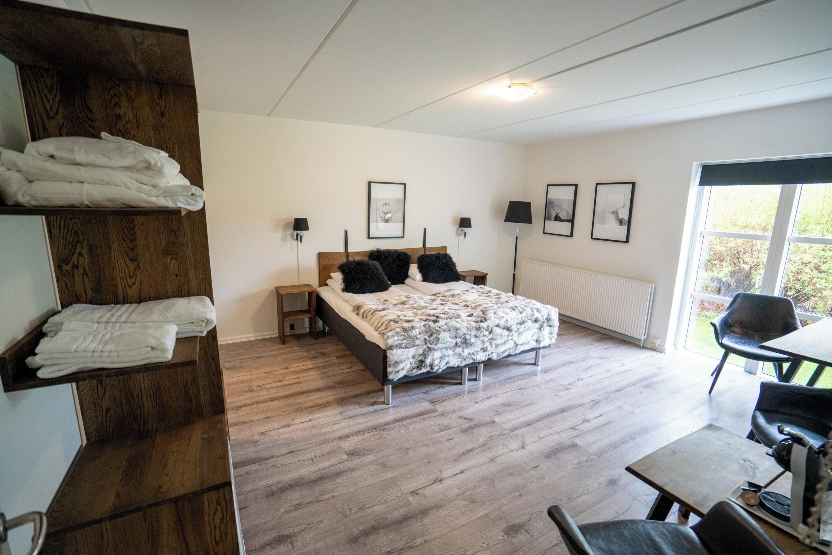 Quarto com cama, mesas de cabeceira, prateleira de madeira com toalhas, radiador e janela com vista.